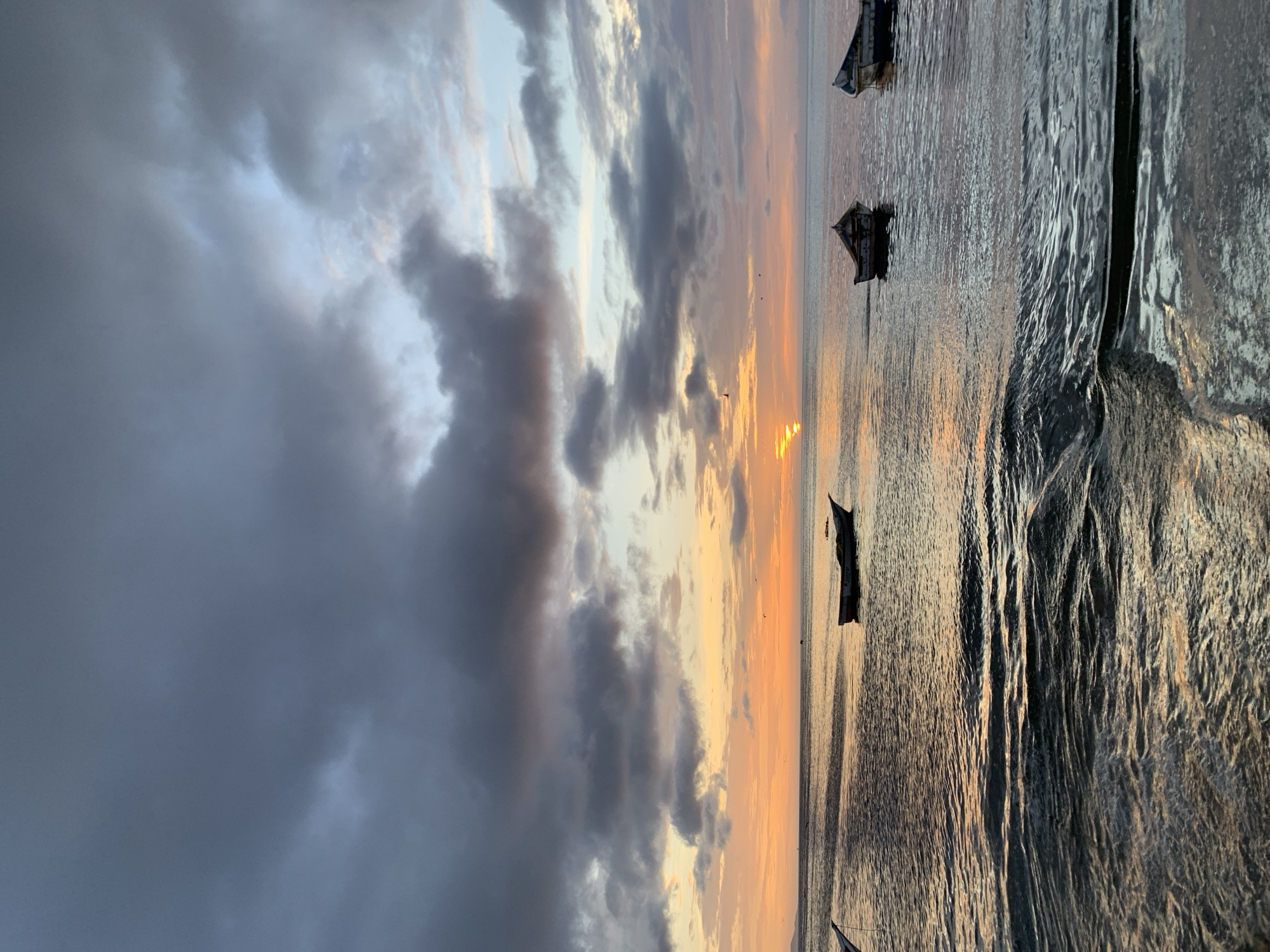 This image captures a serene sunset scene in Juan Griego, Venezuela. The sky is adorned with a dramatic display of clouds, ranging from dark grey to lighter shades of blue and pink, with the setting sun casting a warm orange and yellow glow. The horizon line is clearly defined where the sky meets the calm, reflective ocean. Several small, weathered boats are anchored on the water, their silhouettes stark against the vibrant sunset. One boat in the foreground is partially submerged, adding a sense of age and disuse to the scene. The water itself is rippled, with small waves creating a textured surface that reflects the colors of the sky. The overall impression is one of tranquility and natural beauty.