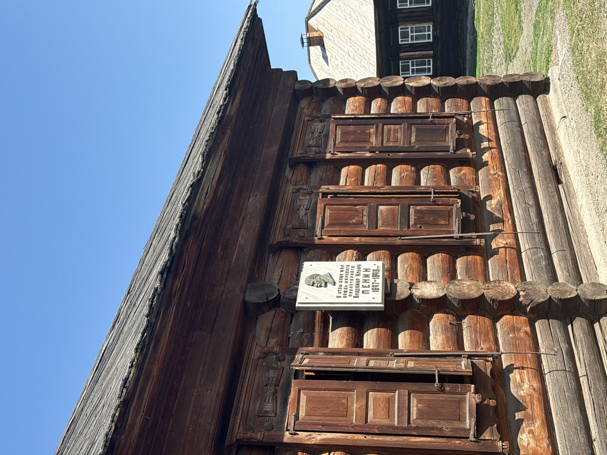 The image shows a historic wooden building in Shushenskoye, Russia, under a clear blue sky. The building is constructed from large, round logs and features several wooden shutters on its windows. A plaque is mounted on the wall, displaying a portrait of a man and text in Cyrillic. The plaque commemorates Vladimir Ilyich Lenin, indicating that he lived in this house from 1897 to 1898. To the right, another wooden building with a gabled roof and white-framed windows is partially visible. The ground in front of the buildings is covered with gravel and grass.