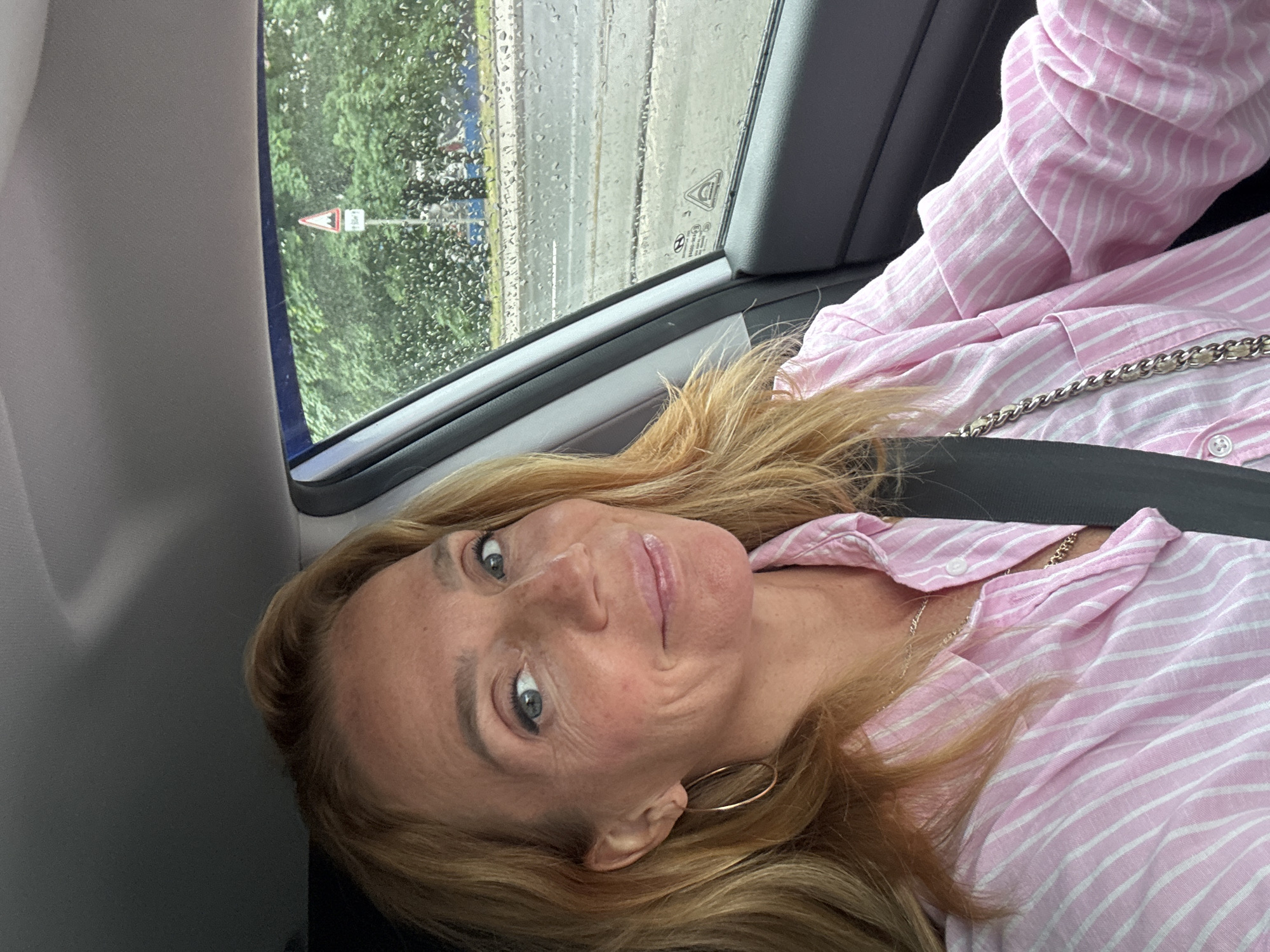 A woman with blonde hair and blue eyes, wearing a pink and white striped shirt, sits in the driver's seat of a car. She is smiling and looking at the camera. Raindrops are visible on the car window, and through the window, a street and trees can be seen. A triangular road sign with a red border and a picture of a sharp curve is visible in the distance. The car's interior is dark gray. The woman is wearing a silver necklace and a large hoop earring in her left ear.
