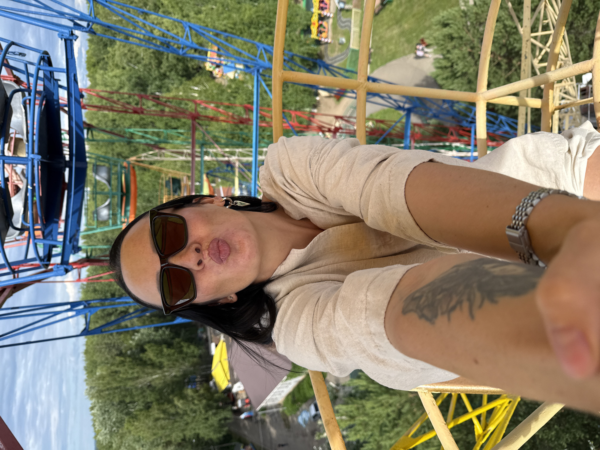 This is a vertical, eye-level selfie taken by a woman in Chaikovsky, Russia. She is in what appears to be an amusement park, possibly on a Ferris wheel or similar ride, indicated by the colorful metal structures in the background and the elevated perspective of the scene.
The woman is wearing sunglasses with dark lenses and a light-colored linen outfit. She is pouting her lips and looking towards the camera. Her hair is dark and appears to be shoulder-length. A silver watch is visible on her right wrist, and a tattoo is visible on her left arm.
The background features a vibrant mix of primary colored metal structures, likely part of amusement park rides, interspersed with lush green trees and a cloudy sky. The lighting suggests it is daytime, with no specific indication of the time of day other than the presence of natural light. The overall mood of the image is casual and lighthearted, fitting for an amusement park outing. There is no visible text in the image.