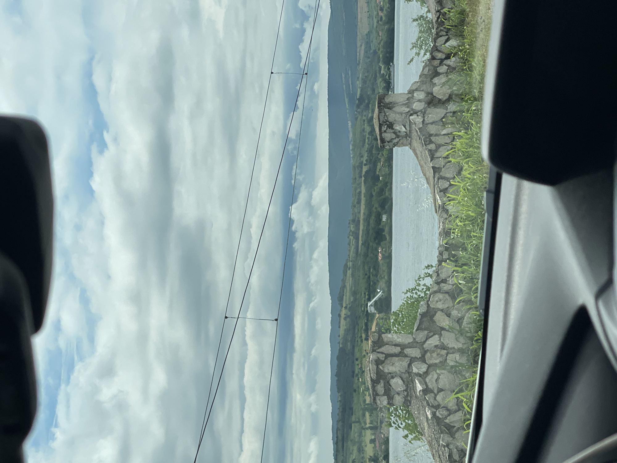 This image captures a wide landscape view, seemingly taken from inside a vehicle looking out through a window. The scene is dominated by a large body of water, distant hills, and a cloudy sky.
In the foreground, elements of a car's interior are visible on both the left and right edges, in dark grey and black tones, framing the picturesque outdoor scene. These include parts of the door frame and window trim, suggesting the photograph was taken from a passenger's perspective.
Immediately beyond the car's interior, a rugged stone wall or embankment runs along the edge of a wide expanse of water, which is light blue-grey and reflects the cloudy sky. Green foliage, including grass and small bushes, grows on and behind this stone structure. A small, light-colored, angular structure, possibly a pier or a small utility building, is visible at the water's edge in the midground.
Stretching across the horizon in the background is a long chain of dark green hills or mountains, appearing to be covered in trees. Above this landscape, the sky is largely filled with white and grey cumulus clouds, with some lighter blue areas visible. Several thin, black utility lines (likely power lines) are strung horizontally across the upper portion of the sky.
The image depicts a serene daytime setting under a cloudy sky, suggesting moderate weather conditions. The presence of a wide body of water and distant hills is consistent with the provided location context of Drobeta-Turnu Severin, Romania, suggesting the water is likely the Danube River. There is no visible text in the image.