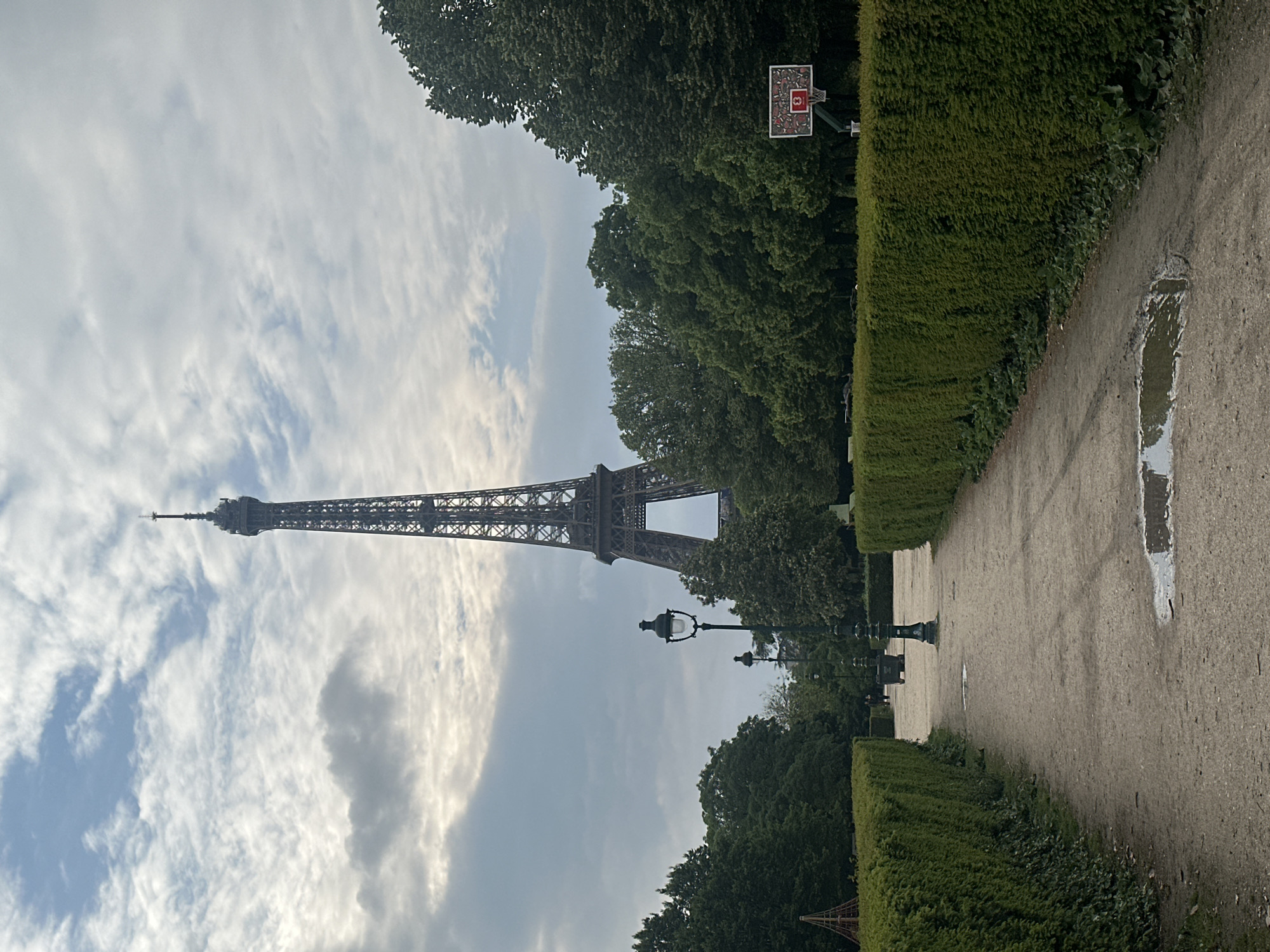 The image depicts the Eiffel Tower in Paris, France, framed by trees and greenery, suggesting a park or garden setting. A gravel path runs along the right side of the frame, bordered by a trimmed hedge. A puddle is visible on the path, reflecting the sky. Streetlamps are positioned along the pathway. In the background, a basketball hoop is visible amidst the trees. The sky is cloudy, with patches of blue.