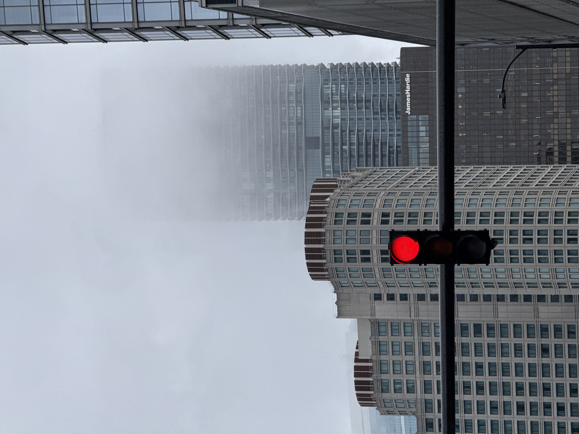 This media file shows a view of the Chicago skyline on a foggy day. The foreground features a traffic light with a red signal illuminated. Behind the traffic light, a light-colored, multi-story building with a repeating pattern of windows is visible. Further in the background, a darker, modern skyscraper with a rippled facade is partially obscured by fog. Another dark-colored building with a grid of windows has the text "JamesHardie" vertically displayed on its side. The overall scene is dominated by a gray, overcast sky and dense fog, suggesting a cool and potentially damp day. The image is oriented vertically, with the bottom of the frame appearing at the top when rotated to a standard horizontal orientation. There are no people visible in the image.