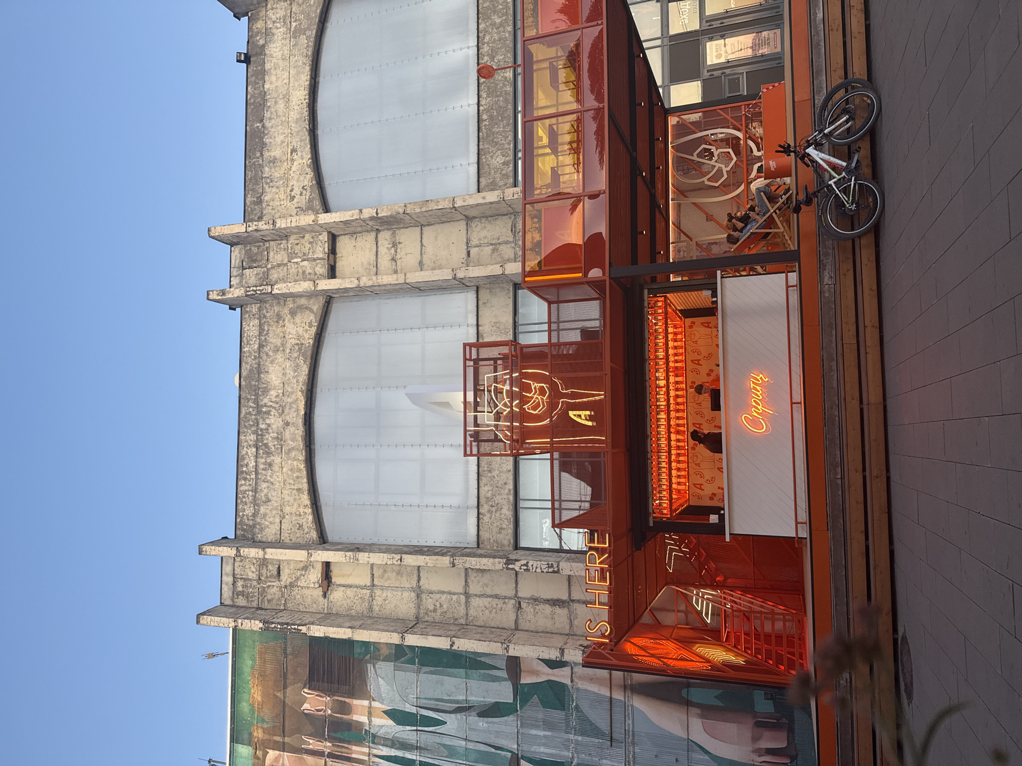 This is a photograph of a modern open-air establishment with bright orange accents, situated in front of a large, industrial-style building. The establishment appears to be a bar or cafe, with a prominent counter and seating areas. Neon signs, one reading "IS HERE" and another in Cyrillic script which translates to "Spritz," are visible.
The building behind the establishment has a weathered facade with large, arched windows that are opaque. To the left of the building, there is a large mural or advertisement featuring a stylized depiction of a hand.
In front of the establishment, on a paved walkway, two bicycles are parked. A few people are seated inside the establishment, some on chairs and one couple lounging in deck chairs. Two individuals are visible behind the counter, possibly staff members. The sky is clear and blue, suggesting it is daytime.
The establishment and the adjacent building are located in the Sevkabel Port, a cultural and entertainment complex situated on Vasilyevsky Island in Saint Petersburg, Russia.