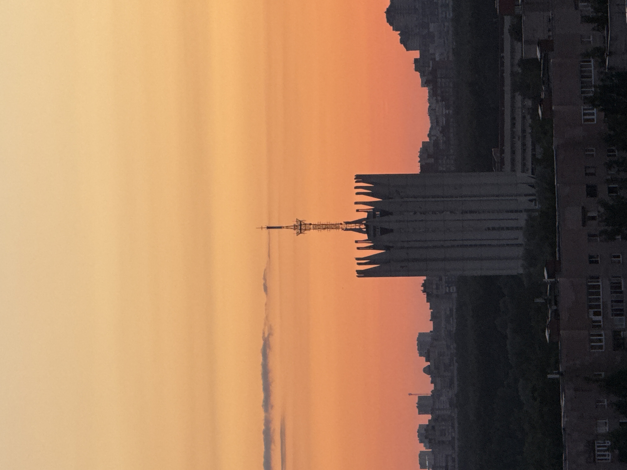The image depicts an urban landscape at either dawn or dusk, characterized by a vibrant, gradient sky transitioning from a light orange-yellow at the top to a deeper orange-red near the horizon. A thin, dark, elongated cloud formation is present in the upper-left quadrant.
The central and most prominent feature is a tall, monumental building in the middleground, constructed from a light grey material, likely concrete. Its architectural style features strong vertical elements extending upwards from the roofline, resembling fins or buttresses. A slender, metallic spire or antenna structure, composed of a lattice framework, rises from the center of the building's roof, culminating in a small, intricate top section.
Behind this central structure, a dark silhouette of a cityscape is visible, comprising multiple high-rise buildings that appear as indistinct blocks against the illuminated sky. A dark band of trees or dense foliage separates these distant buildings from the foreground and the central building.
In the immediate foreground, the darker, shadowed upper sections and rooftops of other urban buildings are visible. These structures display hints of windows and architectural details, but are largely obscured by shadow. No people are discernible in the scene, and no specific actions or interactions are depicted. The scene is set in the city of Unknown, Russia.