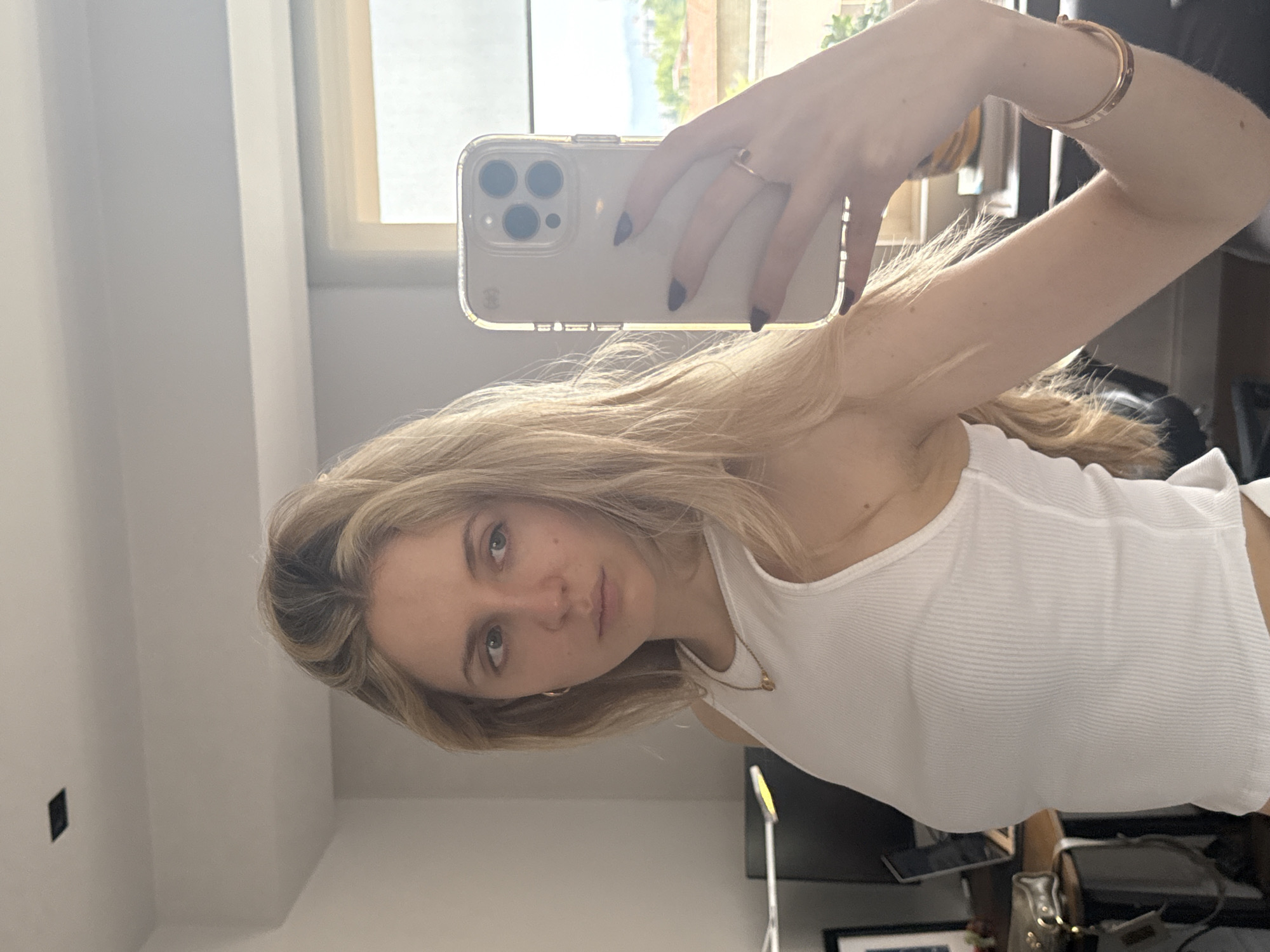 A fair-skinned woman with blonde hair and blue eyes is taking a selfie in what appears to be an indoor setting. She is wearing a white ribbed tank top and a gold bracelet on her left wrist. She also has a gold ring on the ring finger of her left hand. The phone she is holding is white with a clear case. A window is visible in the background, showing an outdoor scene with greenery. The city of Mexico City, Mexico is not discernible from the available details.