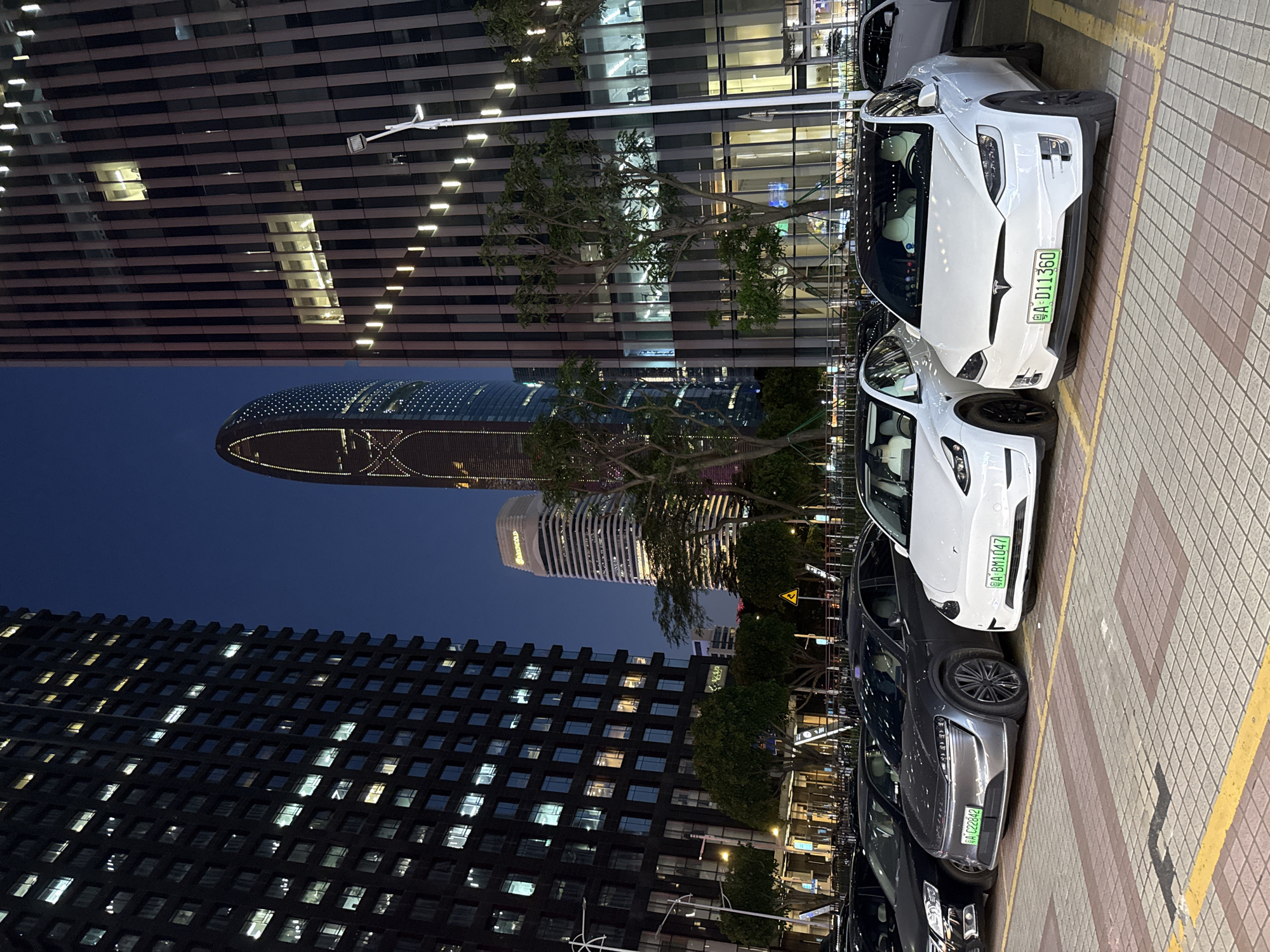 The image captures an urban scene in Tianhe District, China, as indicated by the license plates of the vehicles. The scene is illuminated by the ambient glow of dusk and artificial lighting from surrounding buildings.
A row of parked electric vehicles occupies the right side of the frame. The cars include Tesla Model X vehicles with green license plates, which denotes new energy vehicles (NEVs) in China. The plates are readable, including "A·D11360" and "A·BM1047" and "A·223BAE".
Behind the vehicles, the background is dominated by modern architectural structures. There is a tall building of dark color with many lighted windows. Additionally, there are two curved buildings behind the row of vehicles. The overall setting suggests a well-developed, business area within Tianhe District.