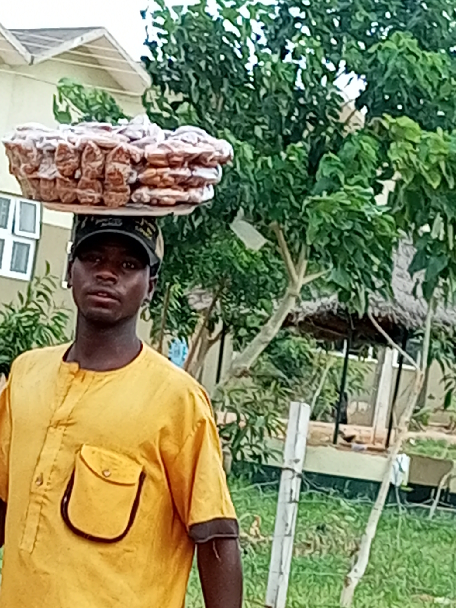 A young man wearing a yellow shirt and a black baseball cap is standing outdoors, balancing a tray of pastries on his head. The pastries are round and stacked several layers high, with a light brown color and a textured surface. The man is looking directly at the camera with a neutral expression. Behind him, there is lush green foliage and a building with a white roof and windows. The environment appears to be an outdoor market or street scene in Gombe, Nigeria. The man is likely a vendor selling his goods.