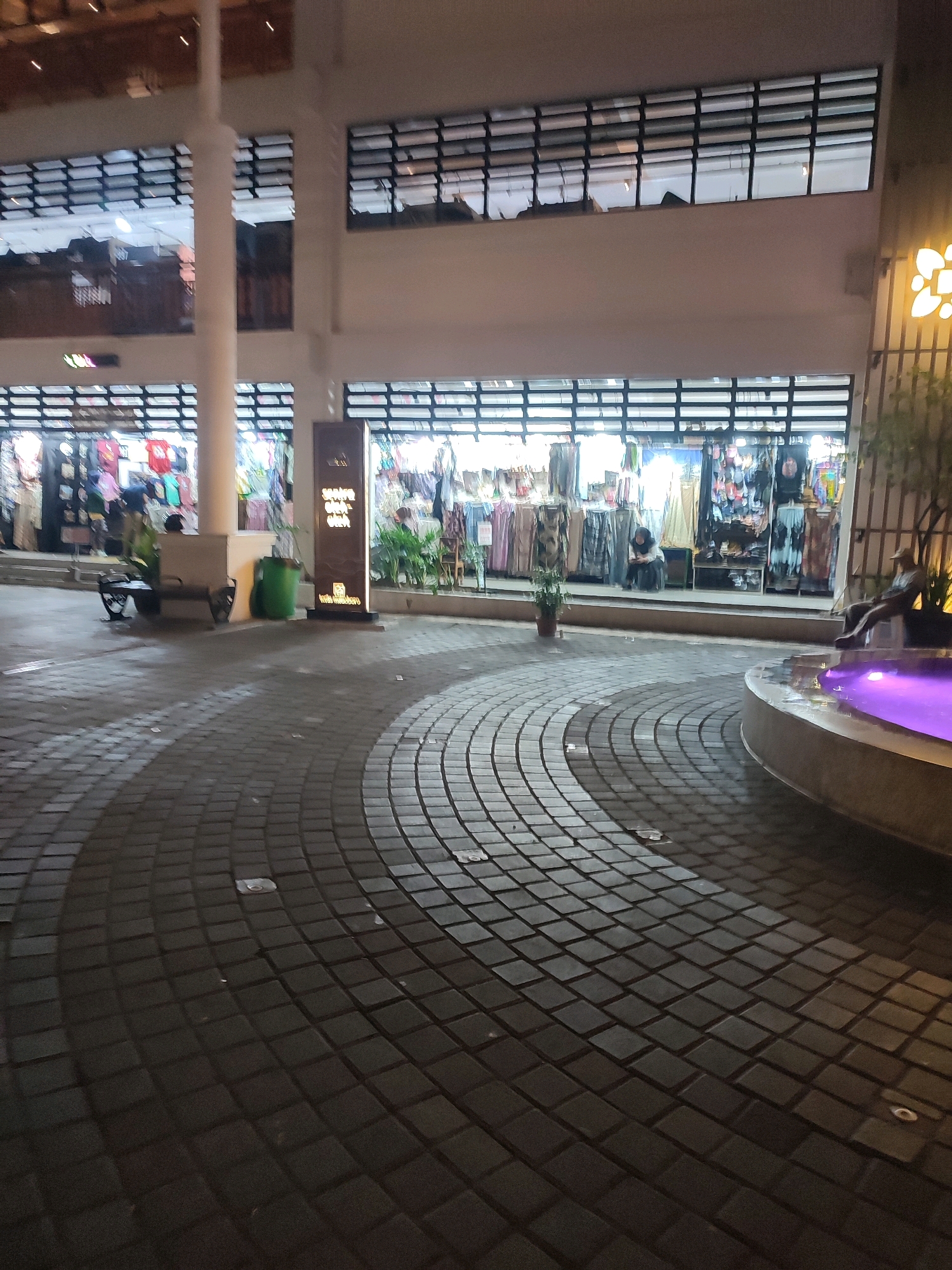 The image depicts a night scene in an outdoor area that appears to be a marketplace or shopping arcade, possibly located in the city of Yogyakarta, Indonesia. The foreground is dominated by a patterned walkway constructed of dark interlocking rectangular pavers, arranged in concentric curves. Lighting from above casts shadows and highlights on the walkway.
In the middle ground, there are several shopfronts with their interiors illuminated. The shops display clothing and other merchandise, with racks of garments visible. One shop has a tall, illuminated sign with text, though it is illegible in the image. To the left of the shops, there is a white pillar, a bench, and a green trash receptacle. A few potted plants are also visible near the shops.
Further in the background, above the shop level, are windows with horizontal louvers, suggesting multiple stories in the building. On the right side of the image, a circular fountain structure is visible, with purple light emanating from the water. A person is seated on the edge of the fountain, and another person is partially visible in the distance, seated on a bench near the shops. The overall atmosphere is that of a commercial area at night, with activity around the shops and a decorative fountain.