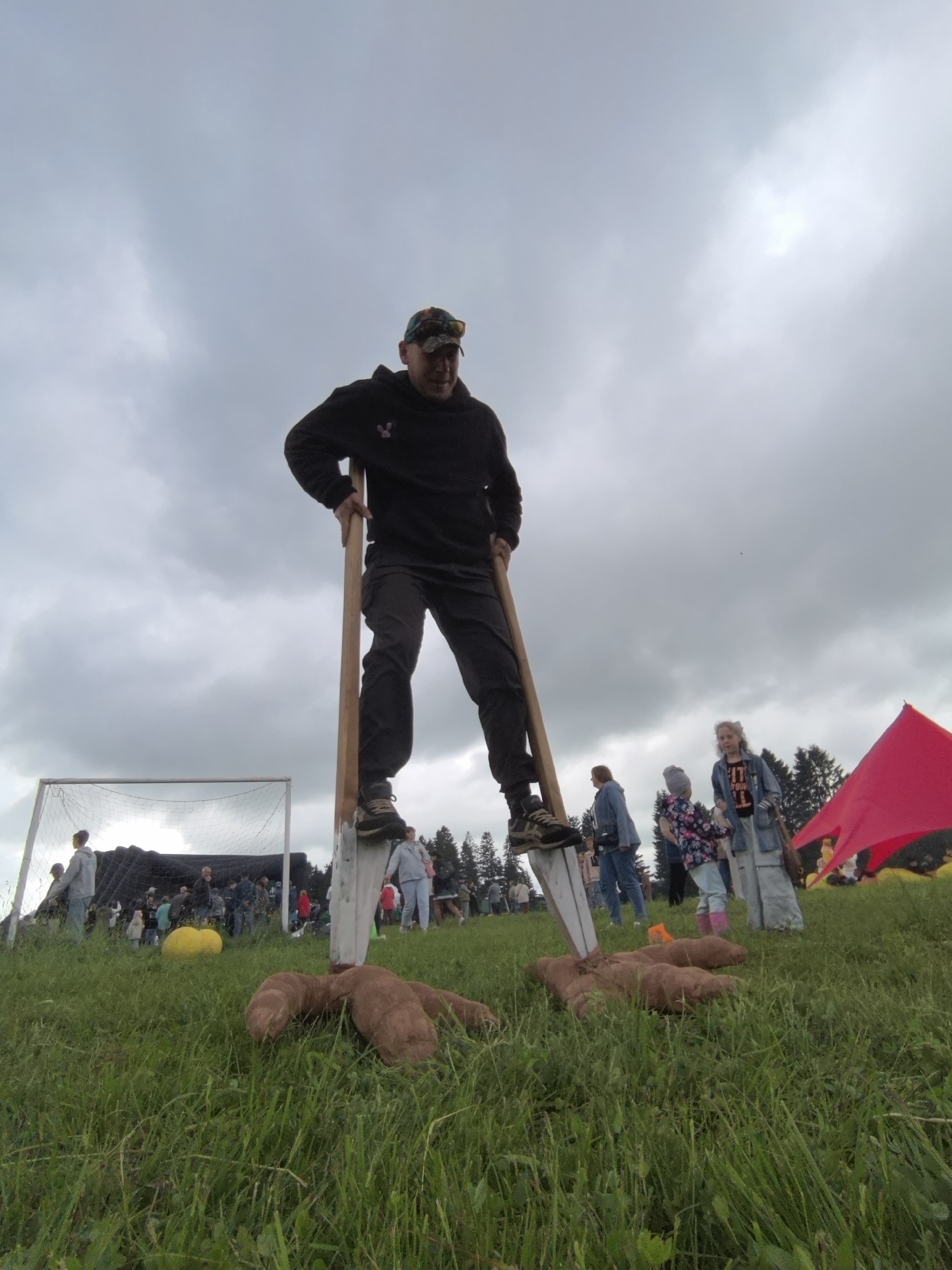 An adult male stands prominently on tall wooden stilts in a grassy field, viewed from a low angle under an overcast sky, in Unknown, Unknown. He wears a black hoodie, dark trousers, athletic shoes, and a camouflage baseball cap with sunglasses pushed up onto the brim, gripping the stilt poles with both hands. Two large, irregular, brown, plush-like objects resembling oversized feet or roots lie on the grass directly in front of the stilts.
In the mid-ground to the left, a white-framed soccer goal with a black net is visible. A large, bright yellow spherical object rests on the grass in front of a crowd of people gathered near a dark tarp or structure. Across the mid-ground and background, a diverse group of individuals, including adults and children, are standing and observing. Noteworthy figures on the right side include a woman in a light blue jacket, a child in a floral jacket with pink boots, and a woman with light hair wearing a denim jacket. A large red fabric structure or tent is partially visible on the far right, nestled among a line of evergreen trees that define the horizon. The sky is uniformly grey and cloudy, indicating diffused lighting over the outdoor scene.