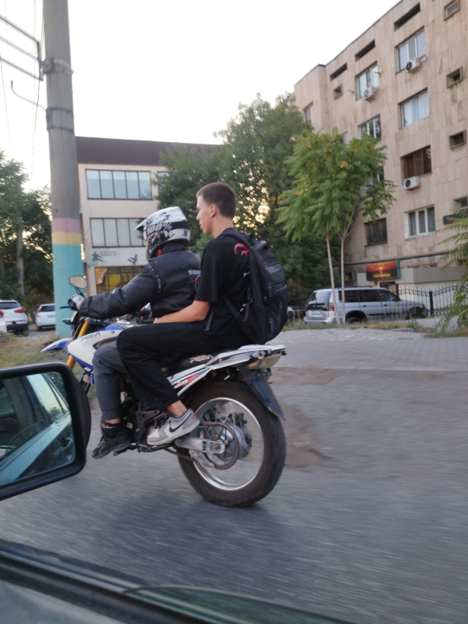 Two individuals are riding a white and blue dirt bike on a paved road. The driver, wearing a white motocross helmet with "100%" branding and a black jacket, is in the front. The passenger, a young male with short brown hair, is seated behind the driver and wearing a black t-shirt and black pants, with a black backpack on his back. He is looking to the right. The dirt bike has "PEDA" written on its side. In the background, a beige apartment building with multiple windows and air conditioning units is visible. To the left of the building, a utility pole with electrical wires stands. Trees with green foliage are also present. Several cars are parked on the side of the road, including a white car, a silver SUV, and a dark-colored vehicle. The scene appears to be in an urban environment in Shymkent, Kazakhstan.
