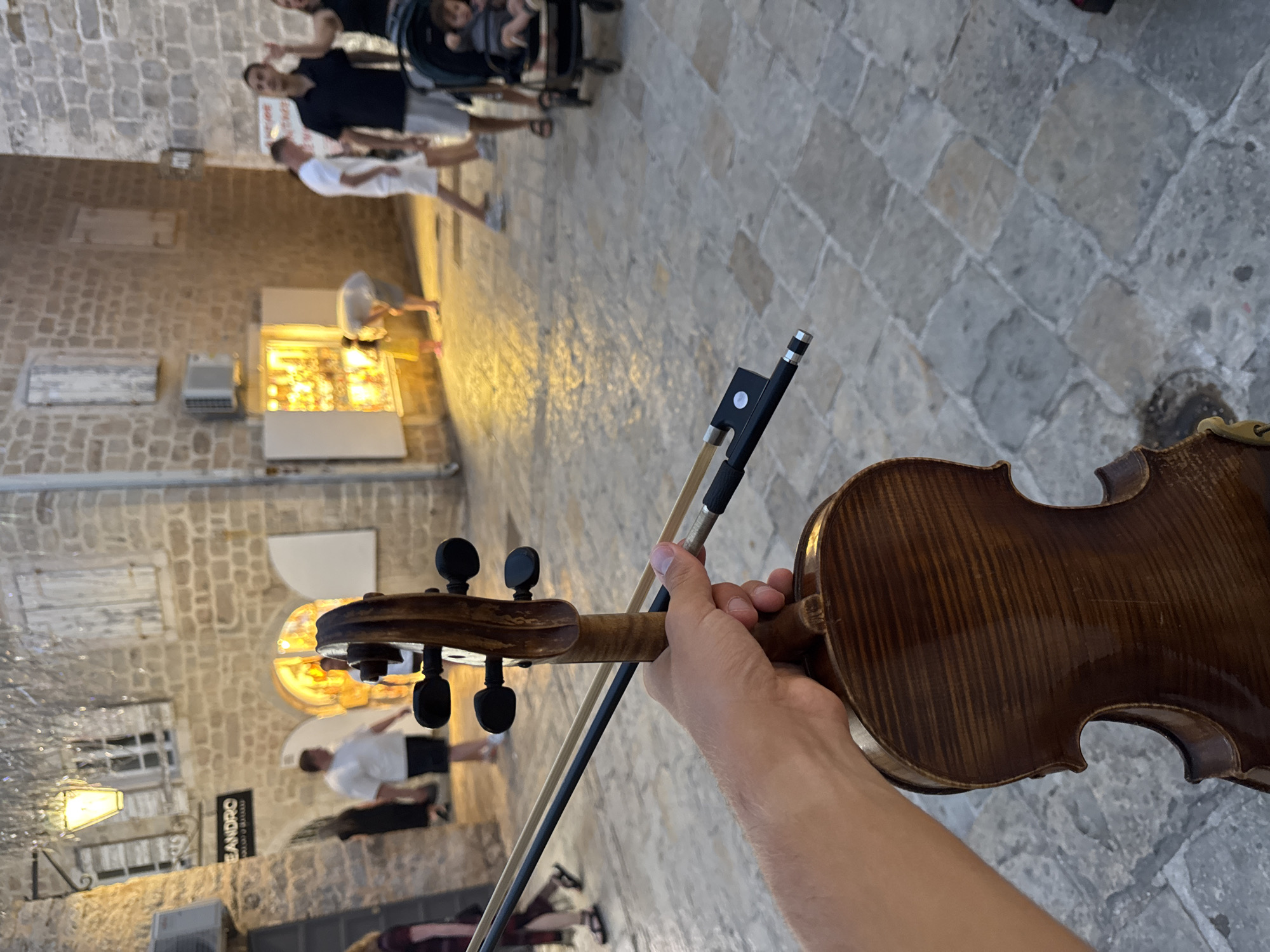 In this image, a person is holding a violin and bow in the foreground, suggesting a musical performance. The background depicts a charming street scene in Budva, Montenegro, characterized by stone buildings with arched entrances and wooden shutters. The street is paved with cobblestones, and there are various shops and lights contributing to a lively atmosphere. Several people are visible in the background, some walking and others gathered near a shop display, including a child in a stroller. The lighting suggests it is either late afternoon or early evening, with warm light emanating from the shops and street lamps. The composition focuses on the violin in the hand, with the blurred street scene serving as a backdrop.