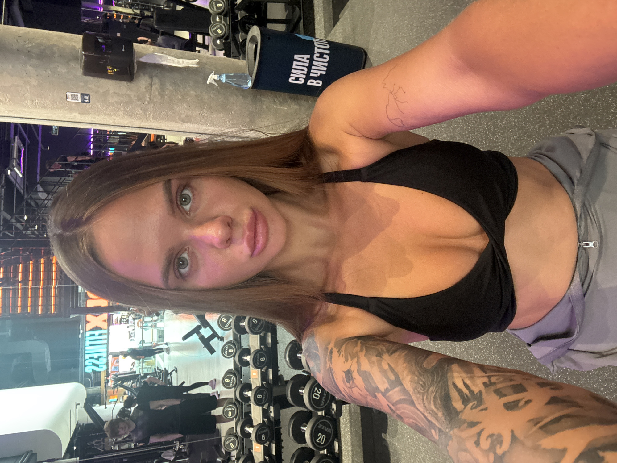 Here is a detailed description of the image:
**General Overview:**
The image is a selfie taken by a woman in a gym. The scene is set indoors, with a combination of gym equipment and design features visible in the background.
**People:**
* **Main Subject:** The primary subject is a woman taking the selfie. She has brown hair and fair skin. She is wearing a black sports bra and grey pants. Tattoos are visible on her arms.
* **Background Figures:** Other people are present in the gym's background, likely working out or using equipment.
**Objects and Environment:**
* **Gym Equipment:** The environment includes various weight training machines, rows of dumbbells, and possibly other types of workout equipment. Dumbbells of various weights are visible.
* **Design Elements:** A gray wall is visible behind the woman. Gym also has a large sign on a wall.
* **Objects:** A trash can with the words "сила в чисто" in Russian on it and a spray bottle are visible.
**Actions and Interactions:**
* **Selfie:** The main action is the woman taking a selfie. Her arm is extended, holding the camera.
* **Workout:** The gym setting implies that individuals are likely engaged in physical exercise or training.
**Location and Context:**
Based on the Cyrillic text ("сила в чисто"), it is likely that the photo was taken in Russia. The text indicates that the trash can says "Strength in Purity". The gym name is "X-FITNESS."
Moscow, Russia.