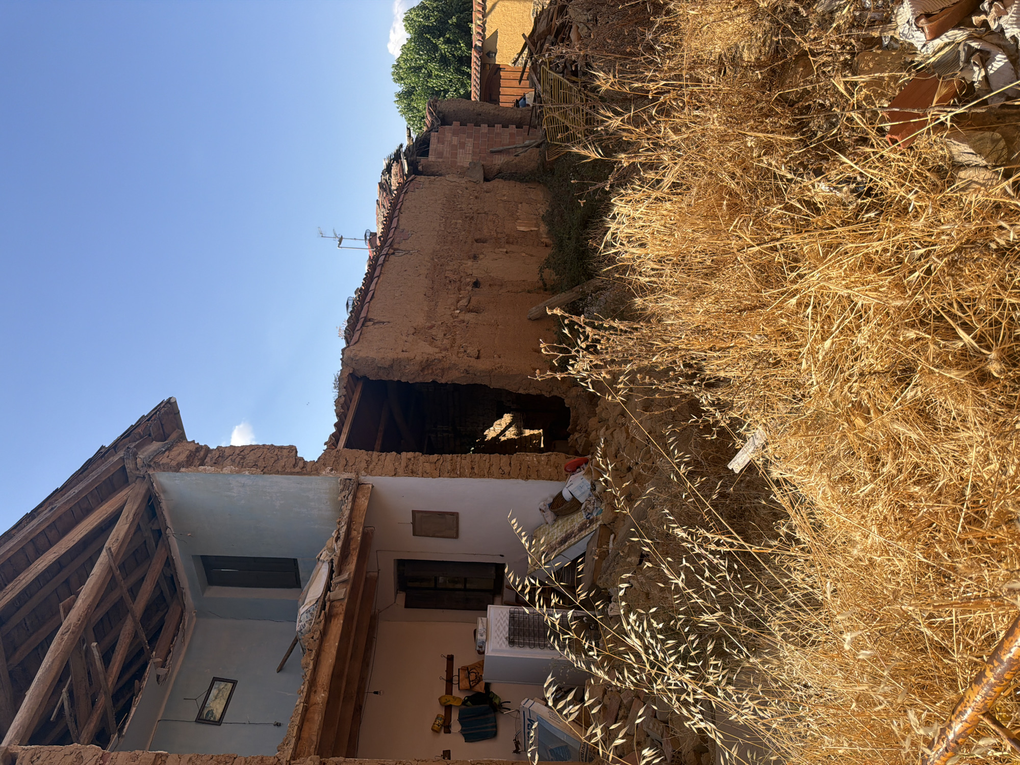 A side view of a damaged, traditional Spanish house in Arcahueja, Spain, under a clear blue sky. The building's adobe walls are crumbling, and the roof has partially collapsed, exposing wooden beams. Debris, including what appears to be a refrigerator and various household items, is scattered around the structure, partially obscured by dry, golden grass in the foreground. The scene depicts a state of disrepair, suggesting it may have been abandoned or damaged by an event. The surrounding environment is rural, with greenery visible in the background.