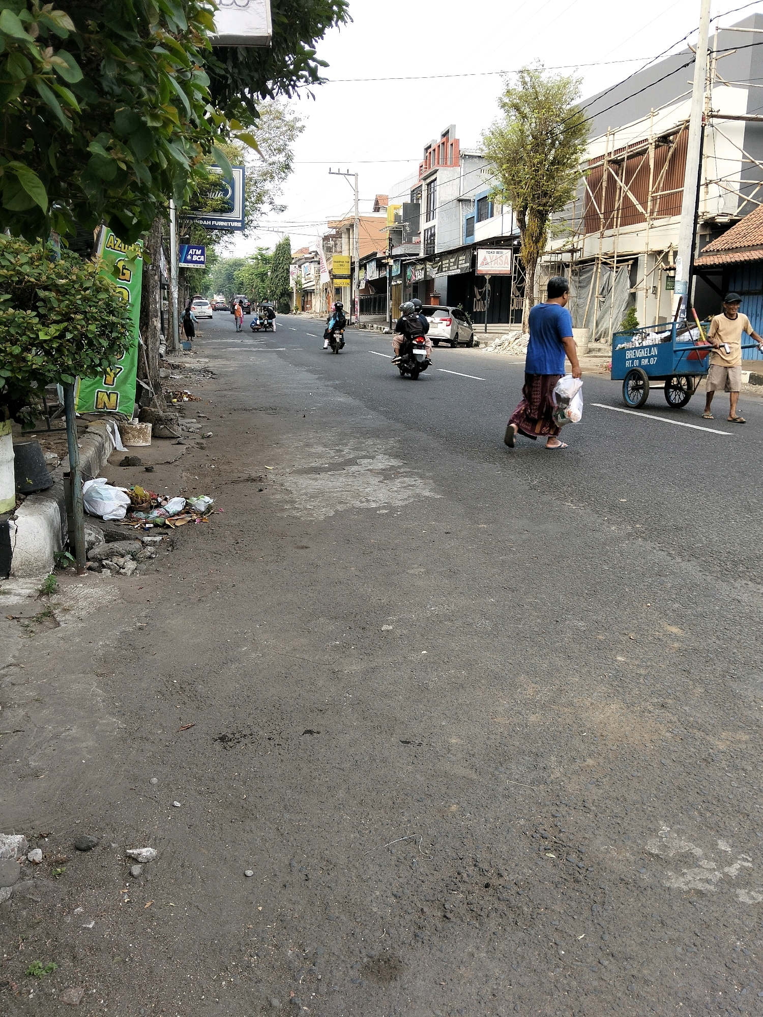 The image depicts a street scene in Purworejo, Indonesia. In the foreground, a pile of scattered trash lies on the side of the road, next to a concrete curb and some greenery. The street itself is paved and appears to be a public thoroughfare with ongoing activity.
Several people are visible in the midground and background. A person in a blue shirt walks across the street carrying a plastic bag. To their right, a man wearing a tan shirt pushes a blue cart. Further down the street, a person on a motorcycle is seen from behind, and in the distance, other vehicles and pedestrians are present.
The buildings lining the street display a mix of architectural styles, with some featuring modern facades and others appearing more traditional. Scaffolding is visible on one of the buildings, suggesting ongoing construction or renovation. The overall atmosphere appears to be that of a busy urban environment. The sky is overcast, casting a diffused light on the scene. The image captures a slice of daily life in this Indonesian city.