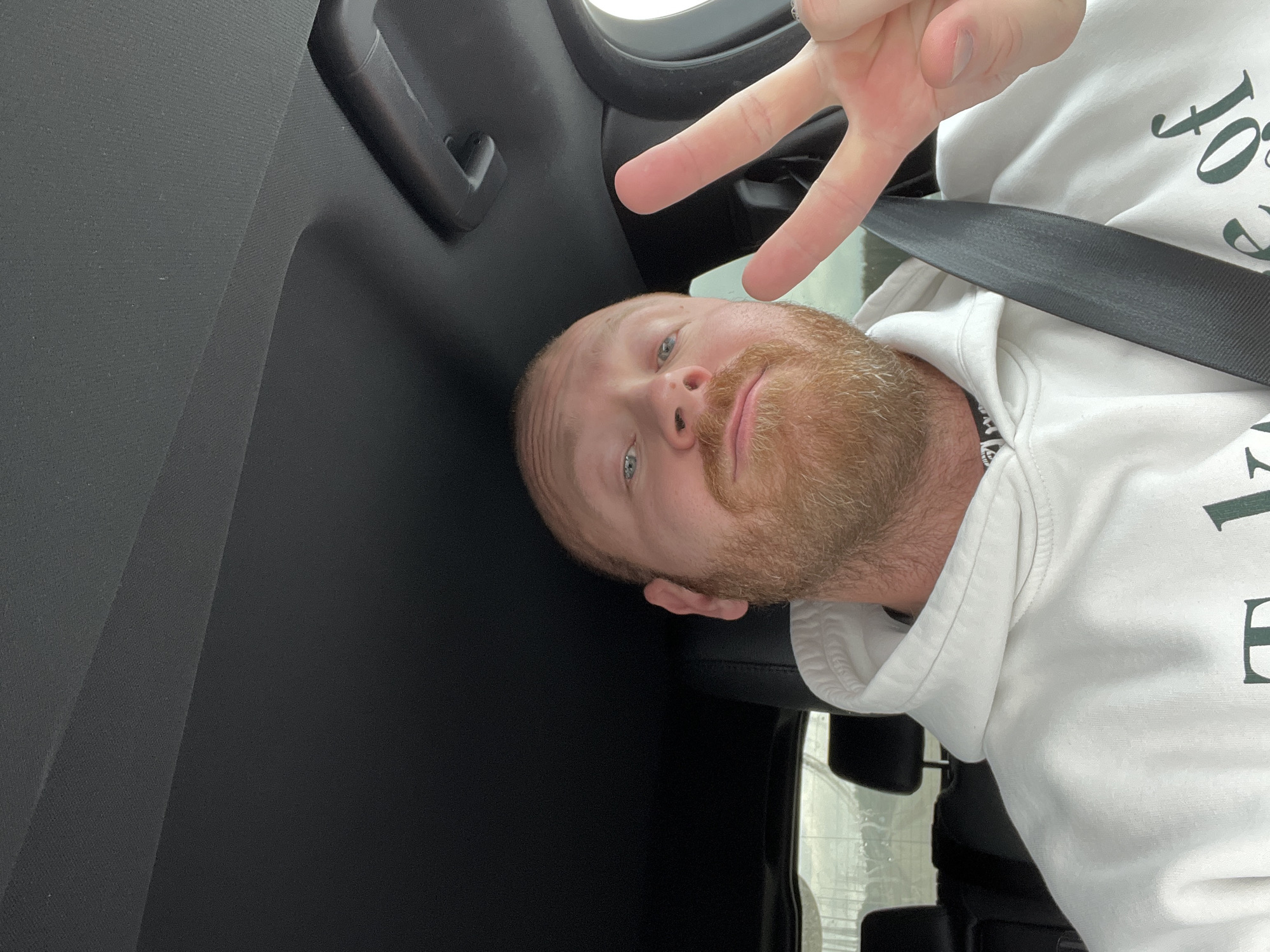 A man with a reddish beard and blue eyes, wearing a white hooded sweatshirt and a seatbelt, is giving a peace sign while seated in a vehicle. The interior of the vehicle appears dark, with a window visible in the background. The location is not discernible as Noviy, Russia, from the available visual information.