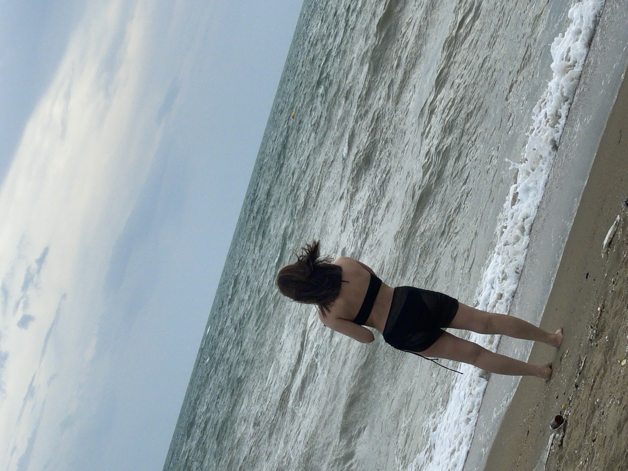 The image depicts a person from behind, wearing a black bikini, standing on a sandy beach. The person's hair is dark and windswept. They are facing the sea, which is choppy with waves. The sky is overcast with clouds. In the foreground, on the sand, there are some debris, including a plastic bottle and a piece of wood. The overall scene appears to be a typical beach environment. There is no specific indication that this location is in Pattaya City, Thailand.