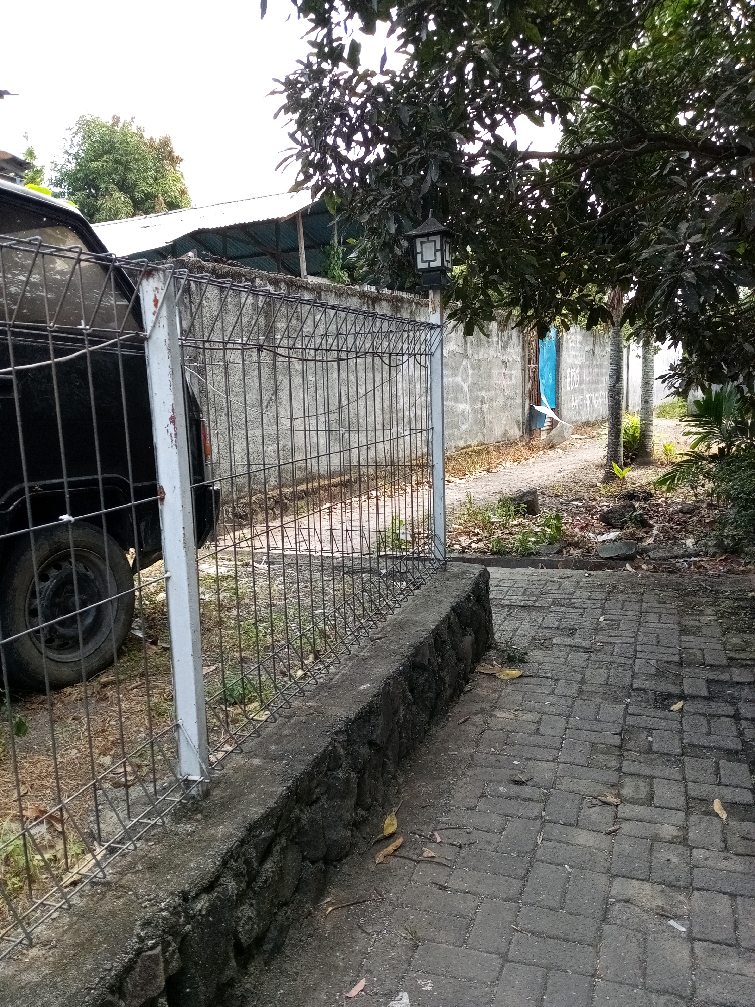 The image shows a view of a fenced-in area with a paved walkway in the foreground, leading towards a wall and a dirt path beyond. A black vehicle is partially visible behind a wire fence on the left side of the frame. The fence is supported by white posts and runs along a low stone wall. A metal lantern is mounted on a post near the wall. Trees with dense foliage overhang the scene from the right and top. The wall behind the fence is made of rough, grey concrete blocks. Further back, a building with a corrugated metal roof is visible. The ground is covered with fallen leaves and debris. The paving stones are rectangular and arranged in a herringbone pattern. The overall impression is of an outdoor area, possibly a yard or a side alley, in a tropical climate, which is consistent with the location of Bitung, Indonesia. There are no people or discernible activity depicted in the image.