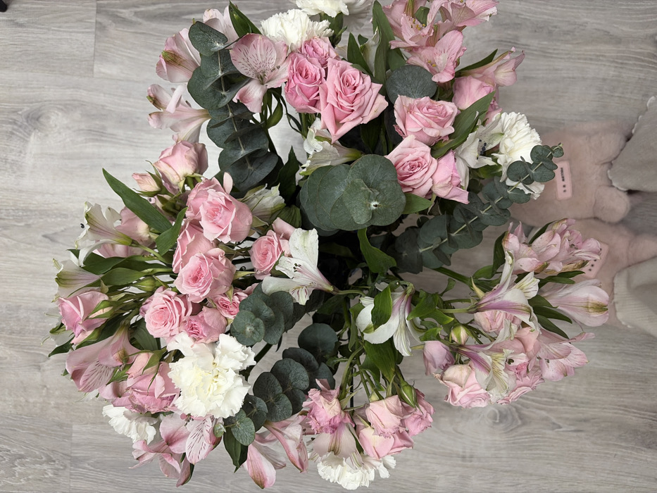 An overhead, close-up shot captures a vibrant floral bouquet resting on a light gray wooden floor. The bouquet is primarily composed of various shades of pink, white, and green.
The main flowers visible are numerous pink roses, ranging from tight buds to partially open blooms, providing a lush, romantic feel. Interspersed among the roses are delicate pink and white alstroemeria (Peruvian lilies), characterized by their striped petals and often speckled centers. Fluffy, ruffled white flowers, resembling carnations or similar blossoms, add volume and brightness to the arrangement. Dark green, rounded eucalyptus leaves are strategically placed throughout the bouquet, offering a contrasting texture and a touch of deep green amidst the softer colors.
The background is a light gray wooden floor with distinct plank lines and a visible wood grain pattern. To the right side of the image, peeking out from behind the flowers, is a soft, light pink, furry or plush object, possibly a slipper or part of a cozy garment. A small tag on this pink object is visible, displaying some dark text that appears to read "R. MATTHEW".
The lighting is soft and even, highlighting the colors and textures of the flowers. The bouquet appears fresh and full, though some of the alstroemeria and lower rose petals in the bottom right section show slight signs of wilting. The overall impression is one of beauty and tranquility.