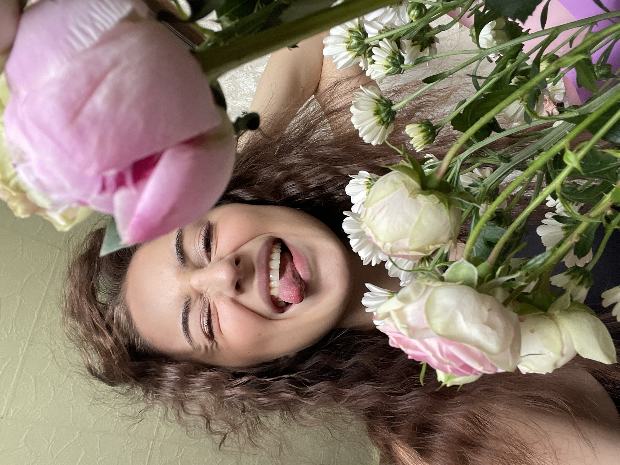A young woman with curly brown hair lies on a light green surface, possibly a bed or couch. She is surrounded by flowers, including a large pink peony on the left and a bouquet of white daisies and pink-tinged white roses on the right. The woman's eyes are closed and she is sticking her tongue out, suggesting a playful or silly pose. The location is not discernible from the image. The image does not include the city of Moscow, Russia.
