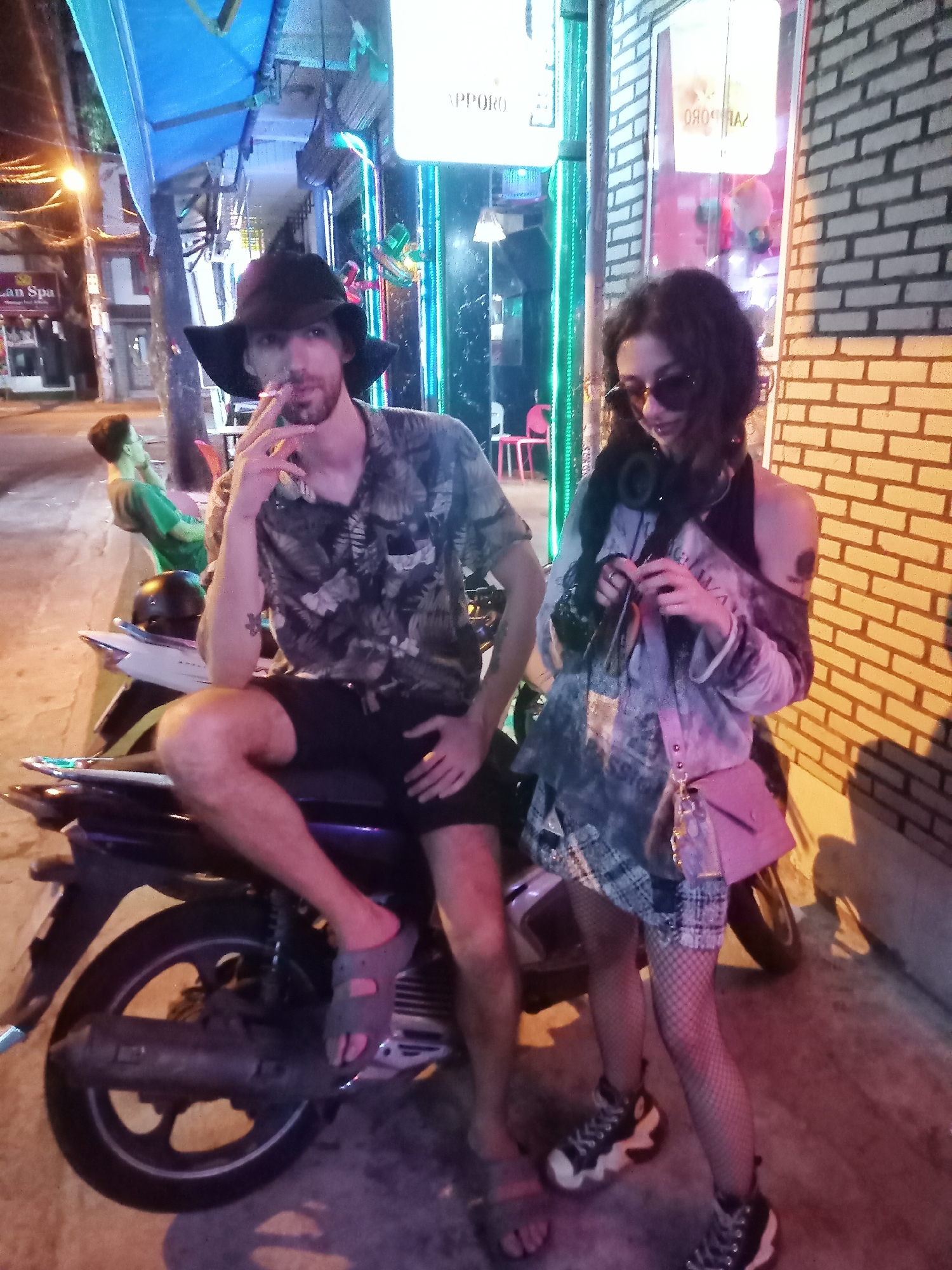 The image captures two individuals on a city street at night, likely in Nha Trang, Vietnam. The scene is illuminated by vibrant artificial lights from streetlights and commercial signs.
The primary subjects are a male and a female. The male is seated casually on a dark-colored scooter, wearing a black wide-brimmed hat, a dark patterned short-sleeved shirt, black shorts, and sandals. He has a beard and is actively smoking a cigarette, holding it to his mouth. A tattoo is visible on his left forearm.
Standing beside the motorbike is the female, dressed in dark sunglasses, a light-colored, long-sleeved distressed top, a plaid skirt, fishnet stockings, and chunky sneakers. She has headphones around her neck and a small pink/purple crossbody bag. She is looking down, seemingly engaged with something in her hands.
In the background on the left, another male figure is visible, sitting on a bench and wearing a green t-shirt. The street features paved ground and brick building facades. Neon lighting and illuminated signs, including a prominent one displaying "SAPPORO" (backwards) and another partially visible as "AN Spa," indicate an urban setting with active nightlife or businesses. The overall atmosphere suggests a casual evening outing.