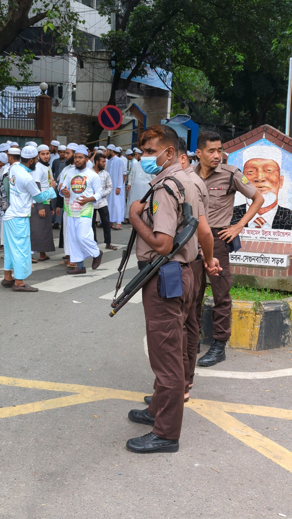 In Dhaka, Bangladesh, two uniformed security personnel stand on a paved street marked with yellow lines. The person in the foreground, facing right, wears a tan uniform with brown pants and black boots, and a light blue surgical mask. They are carrying a shotgun slung over their right shoulder, with a dark blue pouch attached to their belt. Behind them, another security personnel, also in a tan uniform and brown pants, stands with their hands on their hips. In the background, a crowd of men wearing white Islamic attire and caps are visible. A mosaic mural of a man's face is on a brick wall behind the security personnel. The environment appears to be an urban street with trees and buildings in the background.