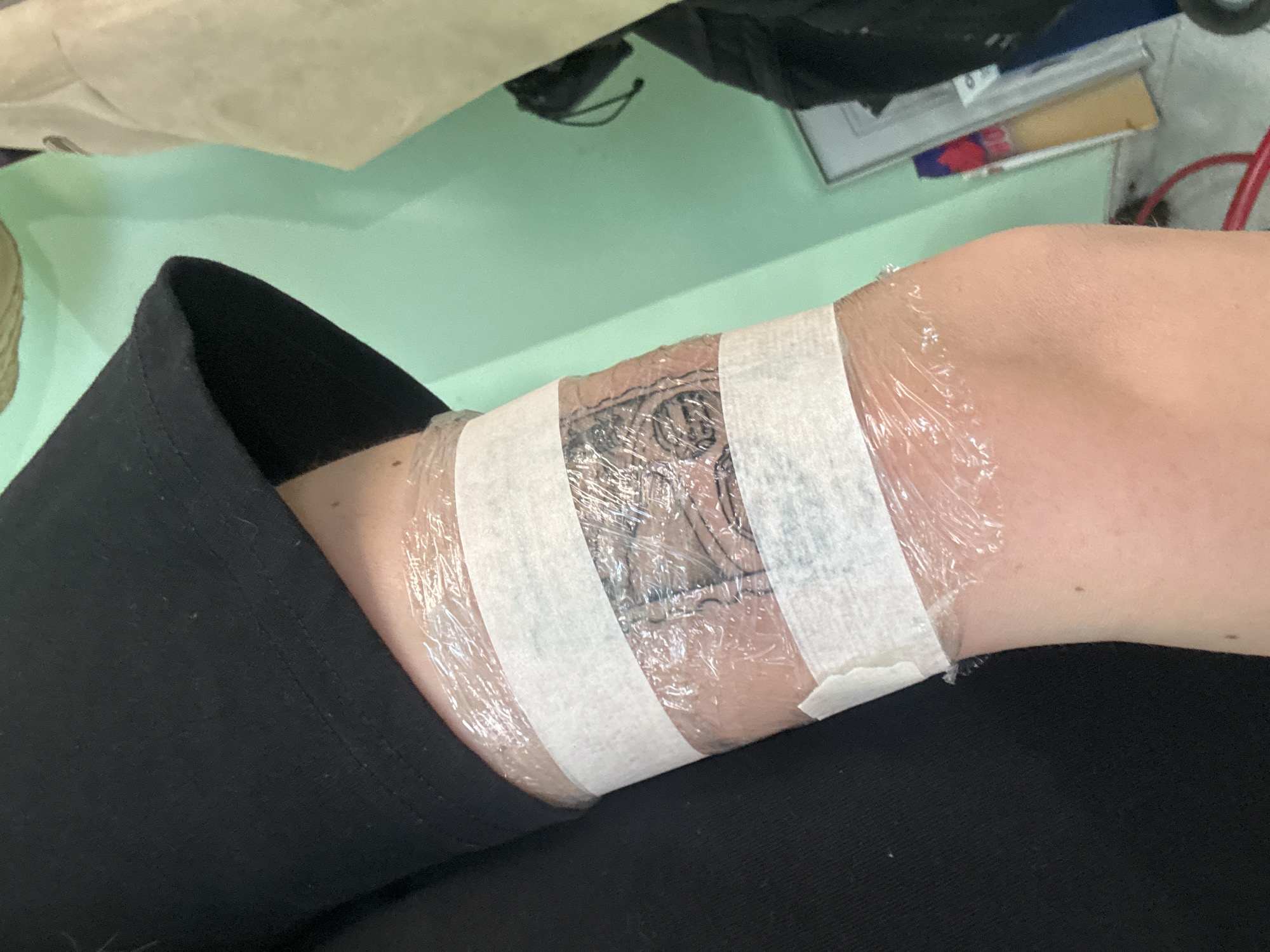 A person's arm is shown with a new tattoo wrapped in clear plastic and held in place by two strips of white medical tape. The person is wearing black pants. The background is a light green surface with some objects partially visible, including what appears to be a book or notepad and some other items. The location cannot be determined from the image alone. There is no information to indicate the city of Varna, Bulgaria.