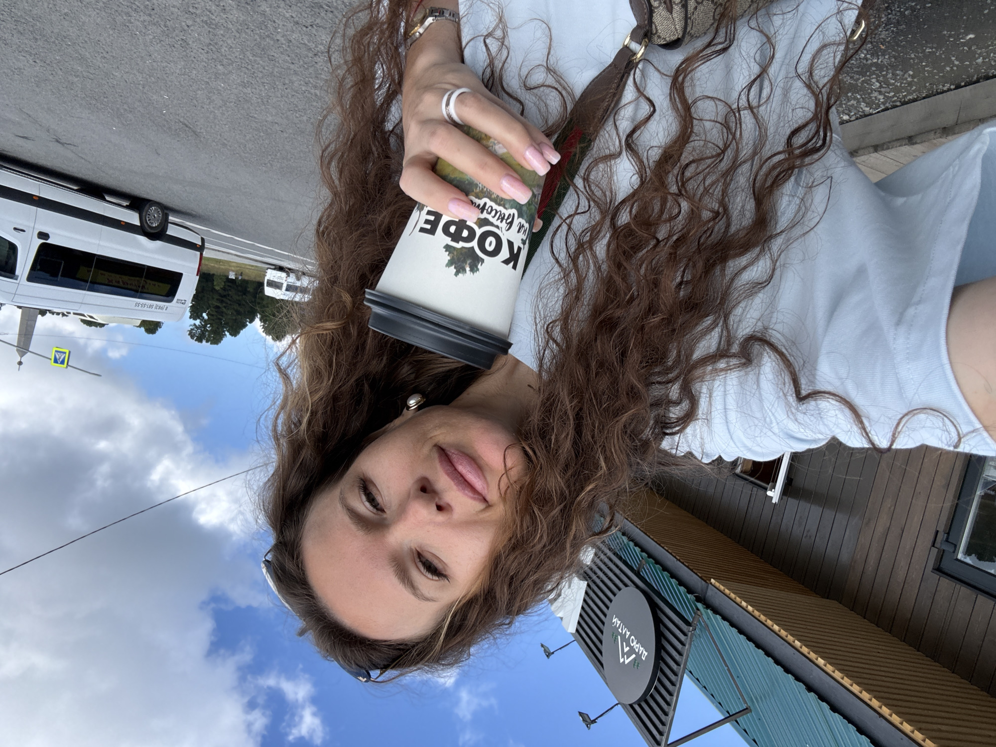 A woman with long, curly brown hair takes a selfie outdoors. She is wearing a light blue t-shirt and holding a coffee cup with Russian text that reads "КОФЕ на высоте" which translates to "Coffee at altitude." The background features a building with a green roof and a sign with the text "ДАРЮ АЛТАЙ" which translates to "Gift of Altai." In the distance, a white van is visible on a road, and the sky is cloudy with patches of blue. The location is identified as T/B Seminskii Pereval, Russia.
