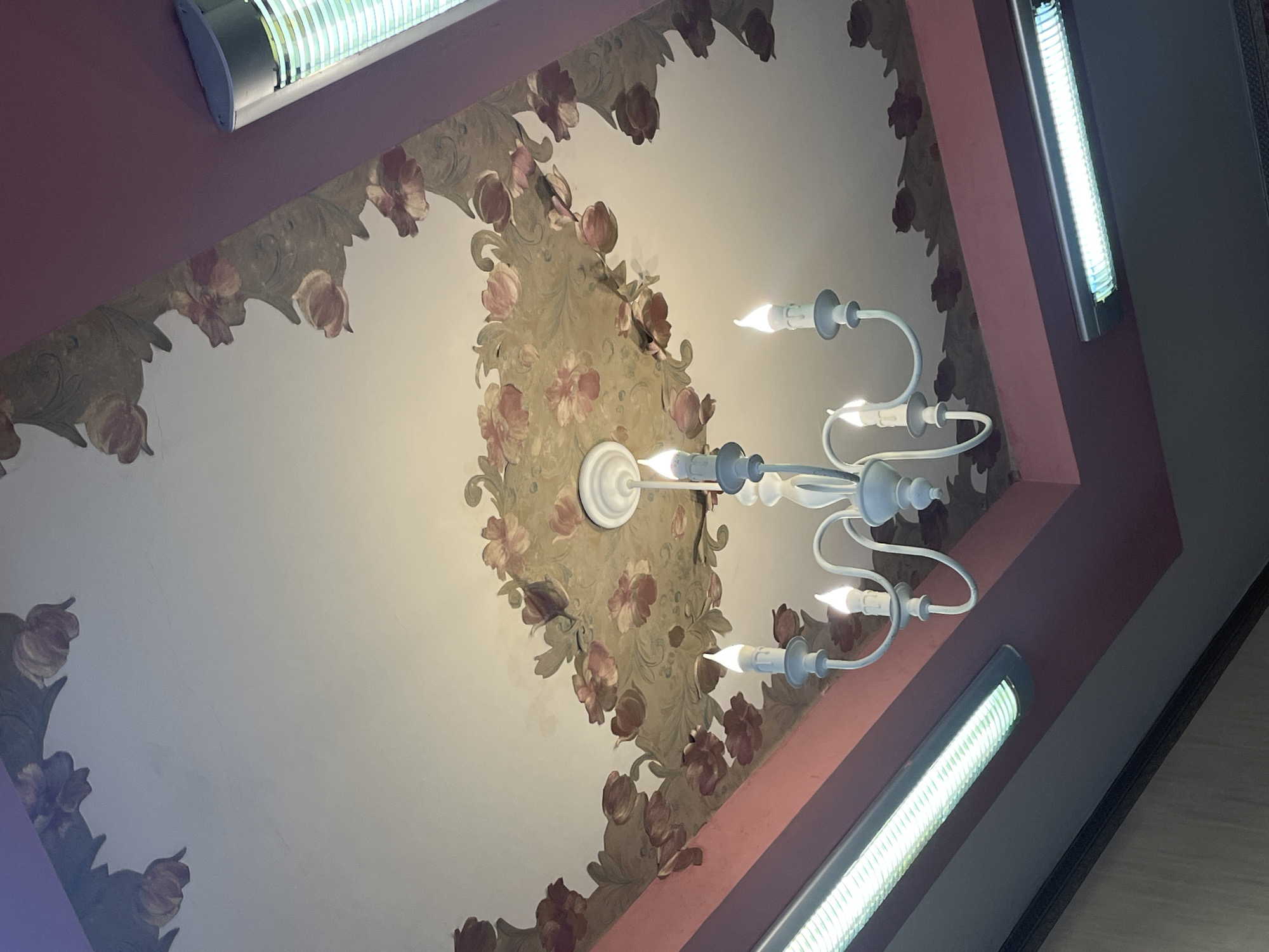 The image displays an ornate indoor ceiling, viewed from a low, upward-angled perspective. A white, traditional-style chandelier serves as a central focal point, featuring multiple curved arms each holding an illuminated, candle-shaped bulb. The ceiling itself is a light cream or white color, adorned with a decorative border and a central medallion around the chandelier. This ornamentation consists of painted or wallpapered floral patterns, characterized by muted green foliage and reddish-pink blossoms, resembling tulips or similar flowers.
Along the periphery of the ceiling, where it meets the wall, two long, rectangular fluorescent light fixtures are visible, emitting a bright, cool white light. One fixture is positioned in the upper left quadrant of the frame, and another is in the lower right, with a third partially visible on the right edge. The walls surrounding the ceiling are painted in a solid, muted reddish-purple hue, which contrasts with the lighter ceiling and some darker, possibly gray or brown, architectural elements on the far right.
No people or visible text are present in the image. The scene is brightly illuminated by the artificial light sources, suggesting an interior setting where external light may not be dominant or it is nighttime. The overall impression is one of an established, decorated interior space in Burgas, Bulgaria.