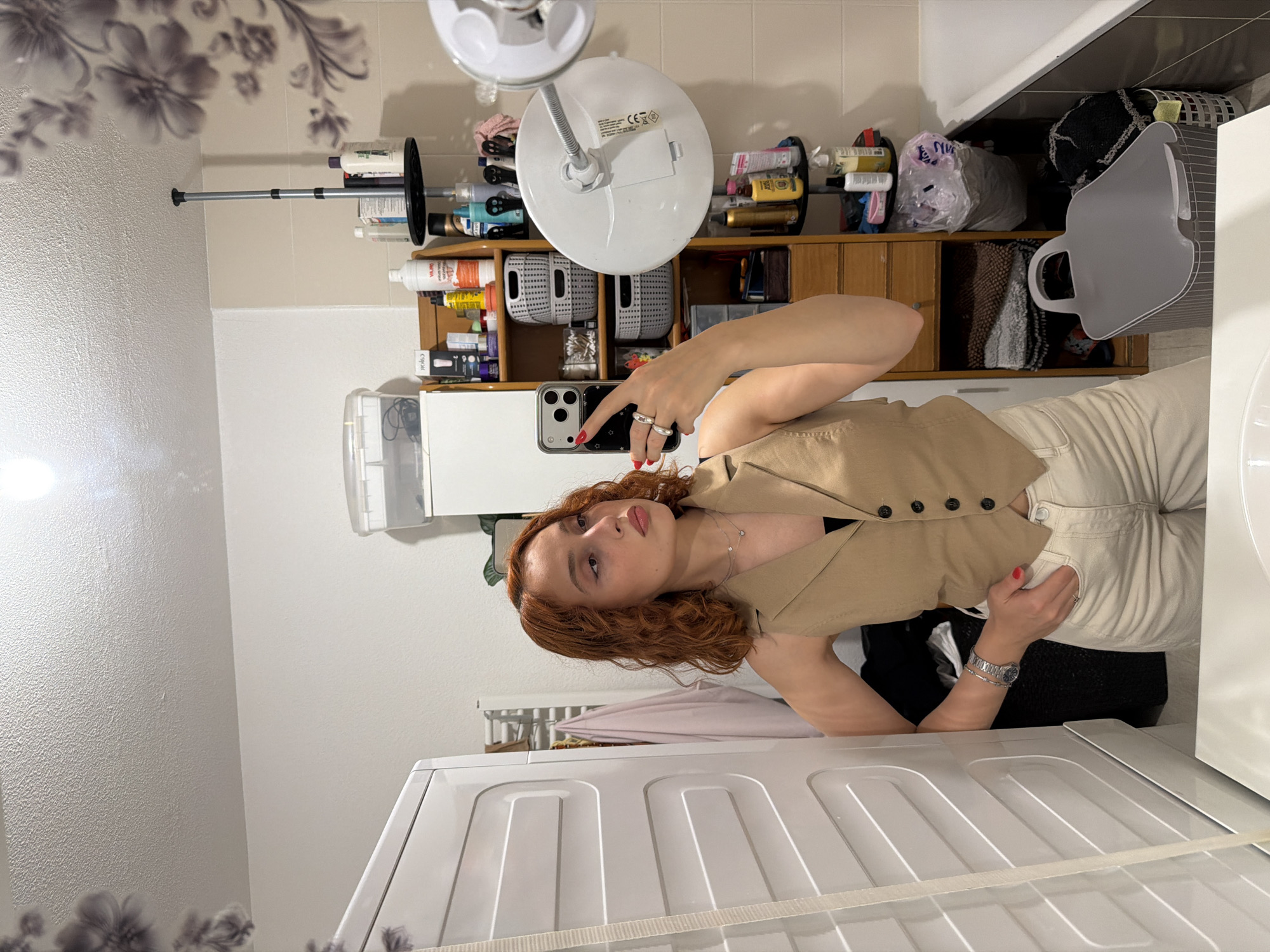 A young woman with curly auburn hair stands in a bathroom, taking a mirror selfie. She is wearing a beige sleeveless vest over a black top and off-white pants. She has rings on her fingers and a watch on her left wrist. The bathroom appears to be in a residential setting, with shelves stocked with toiletries, baskets, and towels. A washing machine is visible on the left side of the frame, and a bathtub is on the right. The overall impression is a casual, everyday moment captured in a private space. The location is not determinable from the image alone.