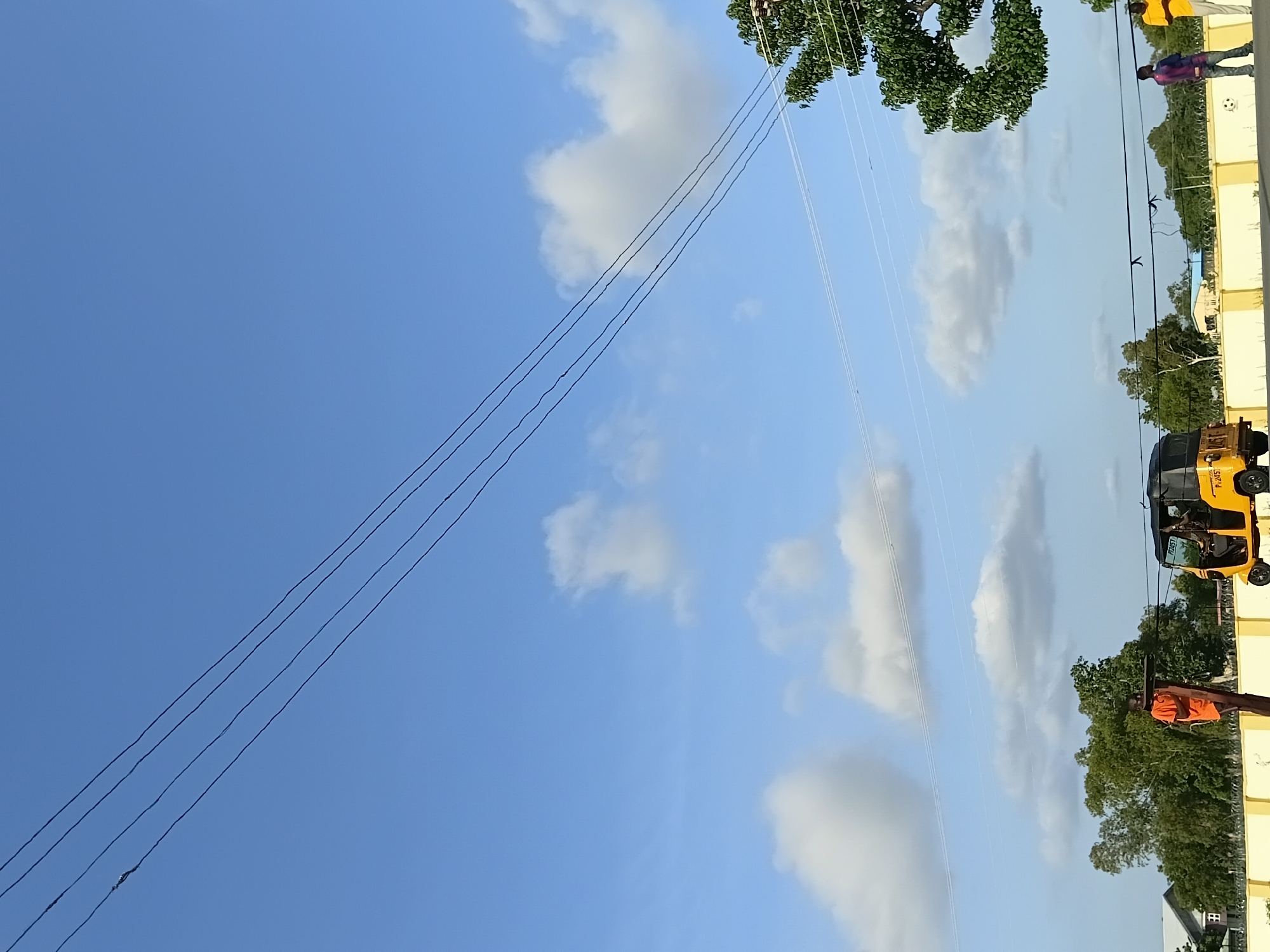 An outdoor scene in Jalingo, Nigeria, shows a clear blue sky with scattered white clouds. Four dark power lines stretch diagonally across the upper portion of the image. In the middle ground, a yellow auto-rickshaw with a black roof is parked. A person wearing an orange shirt stands near a wooden pole on the left side of the image. Further to the right, two men are standing near a yellow barrier fence. The background features lush green trees and some buildings.