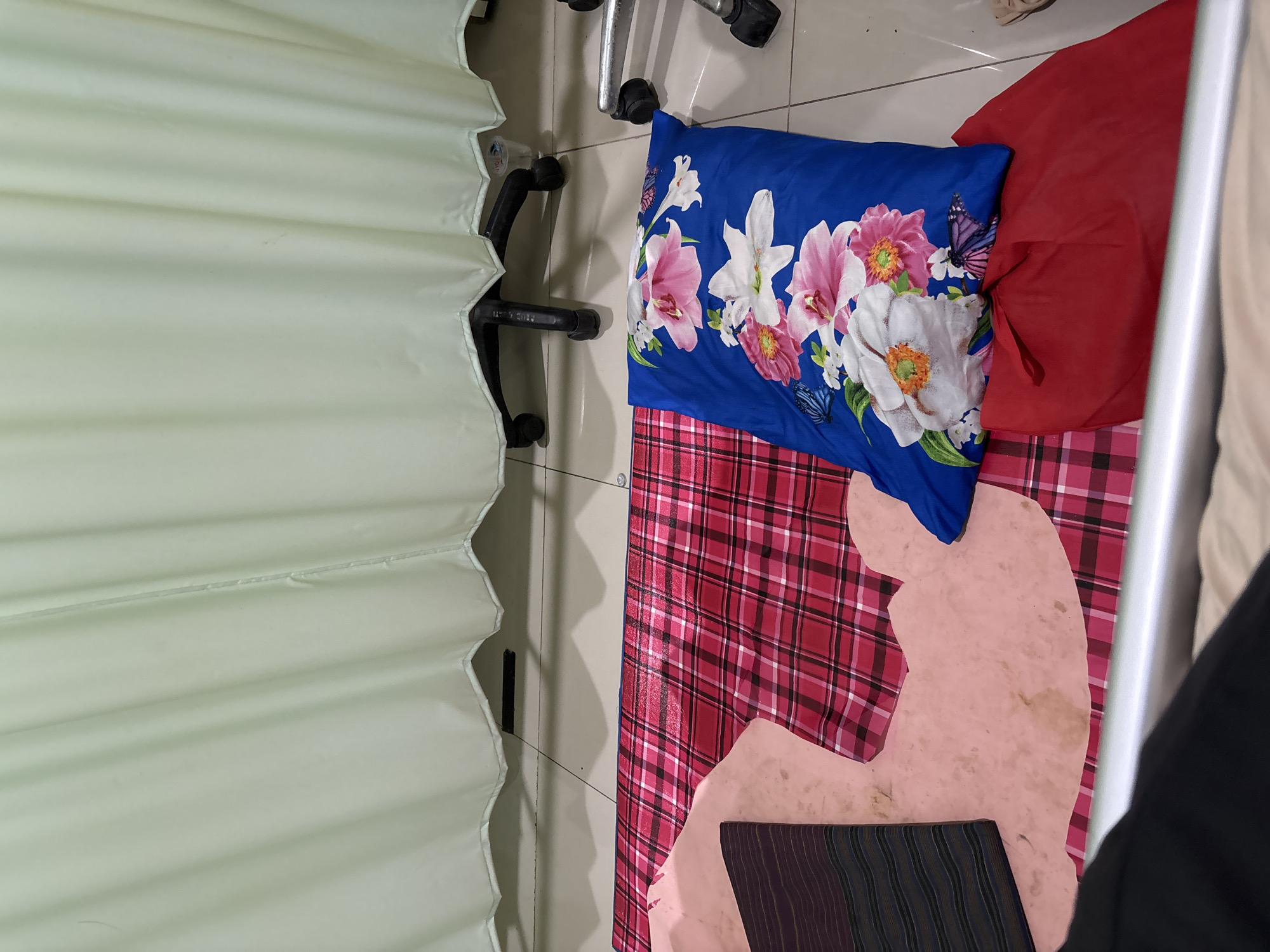 The image shows a scene inside what appears to be a hospital room in Cirebon, Indonesia. A light green curtain hangs in the background, partially obscuring a rolling chair. On the floor, a red and black checkered mat is laid out, with a light pink, oddly shaped material on top of it, possibly a makeshift blanket or padding. A blue pillow with a floral pattern featuring pink and white flowers and butterflies rests on the mat. Next to the pillow is a red bag. In the foreground, a metal railing suggests the presence of a hospital bed. The floor is tiled with a grid pattern.