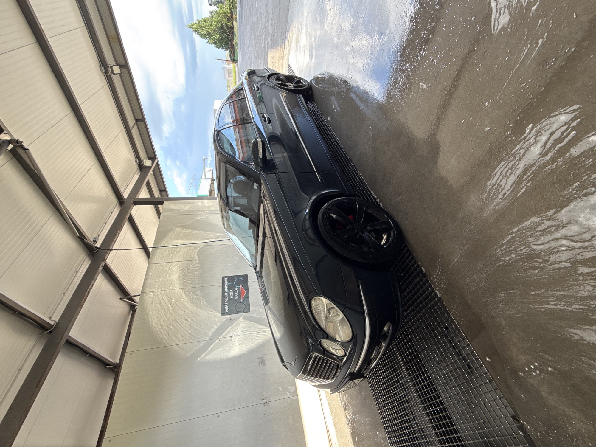 The image shows a dark-colored car, likely a Mercedes-Benz E-Class, parked inside what appears to be a car wash bay in Stara Zagora, Bulgaria. The car is wet and clean, suggesting it has just been washed. The washing bay has a metal grate on the ground, with water and soap suds present. The walls of the bay are made of metal paneling. On one of the walls, there is a sign in Bulgarian that includes the words "пяна ниско налягане вода вакса" (foam, low pressure, water, wax). The lighting is bright, likely due to both natural light and artificial light from the roof. A partial view of the sky and some greenery can be seen in the background, outside the bay.