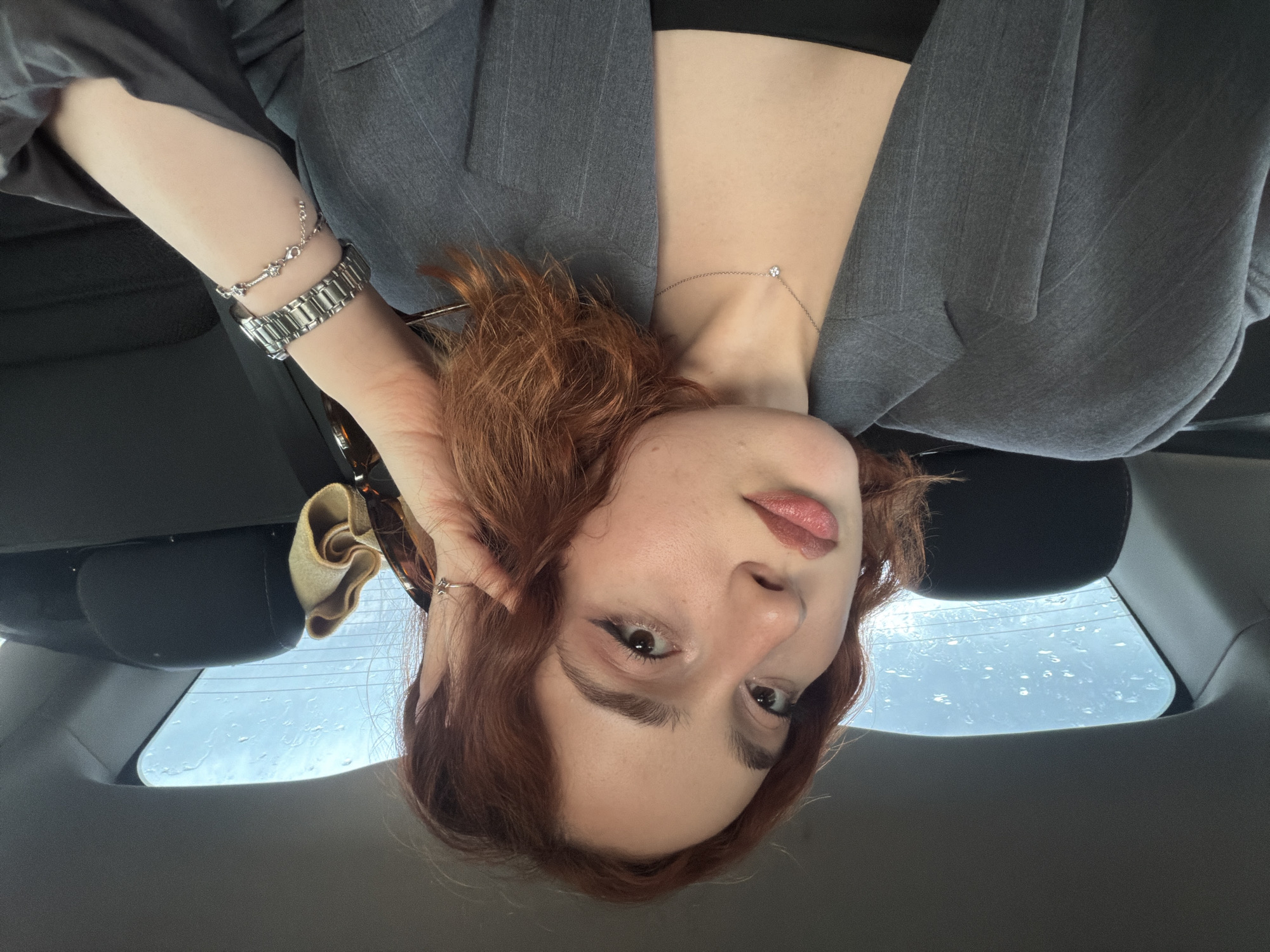 A red-haired woman wearing a grey suit jacket and a black top is in a car in Tbilisi, Georgia. The woman is facing the camera at an upside-down angle. Her left hand is next to her face and she has a watch and a bracelet on her wrist. She is wearing a necklace. The back window of the car is visible, showing raindrops on the glass.