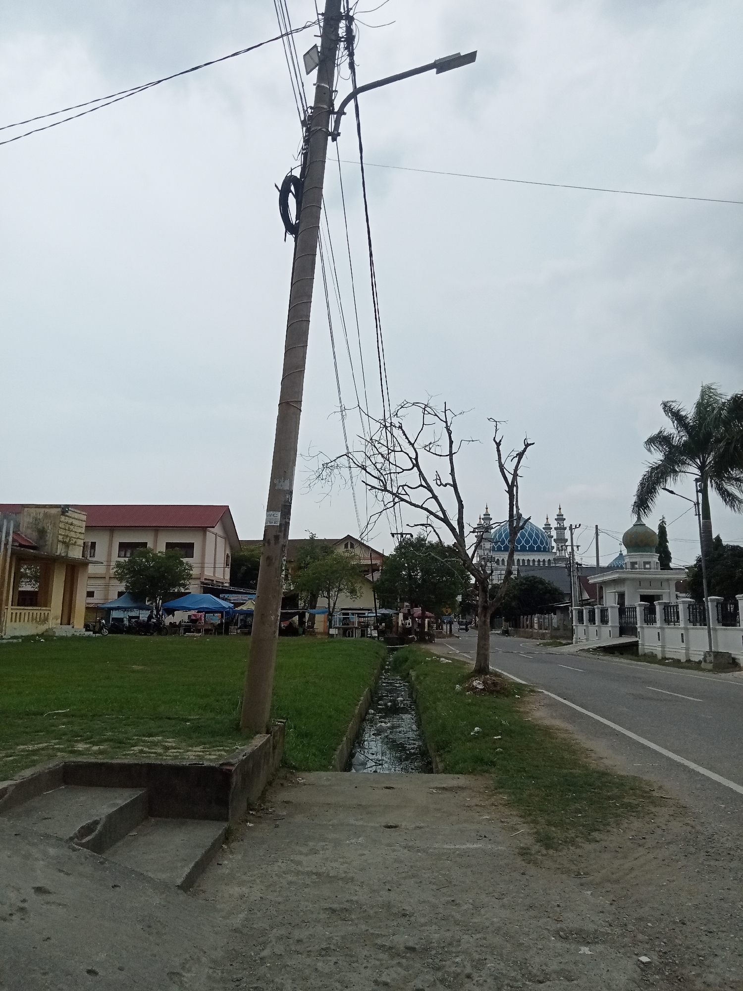 The image depicts a street scene in Kota Lhokseumawe, Indonesia, on an overcast day. In the foreground, a concrete path with steps leads towards a narrow, debris-filled drainage ditch running alongside a grassy area. A thick concrete utility pole stands prominently to the left, supporting multiple electrical wires and a street lamp.
Beyond the ditch, a green grassy field is visible, dotted with several blue and dark blue tents, suggesting a market or an event. Behind the field, a building with a red roof and light-colored walls is situated, to the left of which appears to be another structure with a yellow facade.
Further down the street, a road with a white lane marker curves to the right. On the right side of the road, a white mosque with a green dome and blue domes is visible, along with its entrance gate and white fence. A palm tree stands to the right of the mosque. A leafless tree with a tangled branch structure stands between the foreground and the mosque, on the left side of the road. The sky is uniformly grey and cloudy. No people are clearly discernible in the image, though some objects under the tents might suggest their presence.