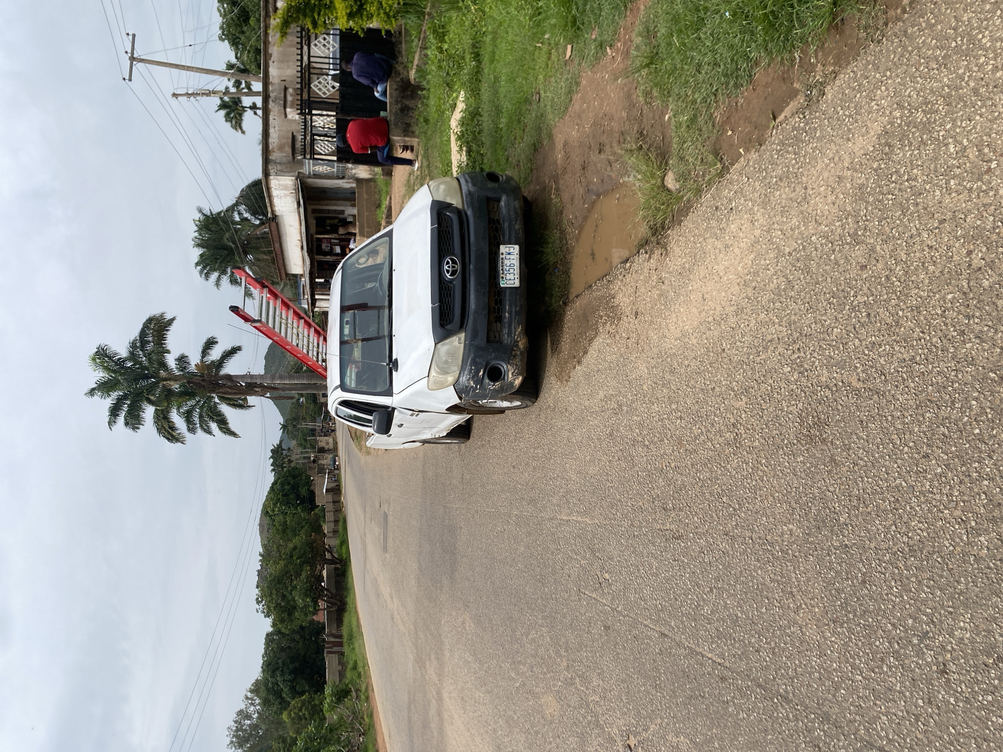 The image shows a white Toyota Hilux pickup truck parked on the side of a paved road. The truck is facing towards the right side of the frame and appears to be a bit dirty. A red ladder is leaning against a building in the background, and several people are visible near the building, some sitting and others standing. The scene appears to be in a somewhat rural or semi-urban setting, with vegetation and trees surrounding the area. There are power lines and poles visible in the background as well. The sky is overcast, suggesting a cloudy day. The license plate of the truck is visible and reads "CC356FKJ". The overall mood of the image is neutral.