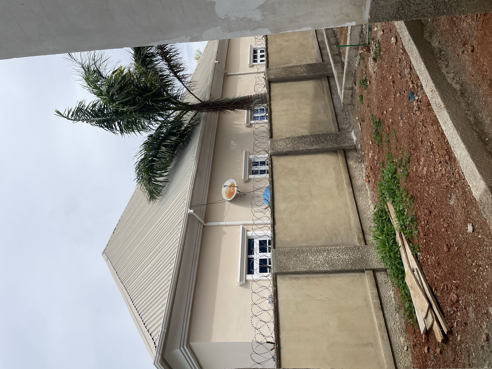The image shows a side view of a tan-colored house with a metal roof in Bar Gada, Nigeria. A tall palm tree stands in front of the house. A concrete wall topped with barbed wire fence encloses the property. The ground in front of the wall is red soil with some scattered wooden planks and green vegetation. Further in the foreground, there are concrete foundations for a new structure. The sky is overcast.