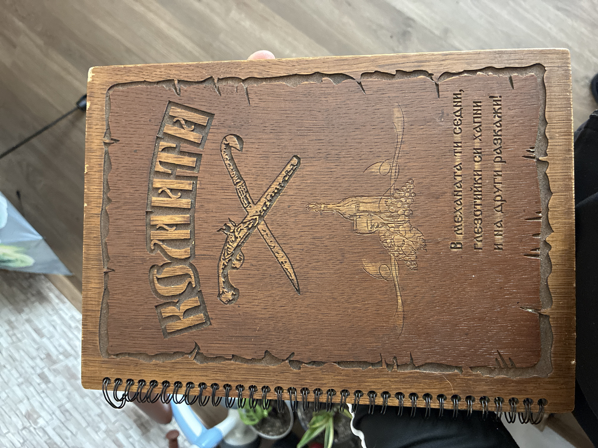 The image displays a close-up, overhead view of a spiral-bound wooden notebook or album. The cover is crafted from dark-stained wood, featuring a rich, carved or engraved design. A roughly textured, torn-paper-like border frames the central design area.
On the left side of the cover, the word "КРОЙКАТА" (KROYKATA) is carved vertically in bold, blocky letters. Centrally located are two engraved items: a pistol crossed with a saber or dagger, both detailed with intricate patterns. To their right, an engraving depicts a sailing ship or boat, alongside a wine bottle and a cluster of grapes, suggesting a theme of maritime adventure and enjoyment.
To the far right, three lines of text are carved vertically in Bulgarian Cyrillic:
"В механата ти седни,
глезoтийки си хапни
и на други разкажи!"
This translates to:
"In the tavern you sit,
eat delicacies
and tell others!"
The notebook is bound by black metal spirals along its bottom edge. The background shows parts of a light-colored wooden floor, various potted plants, and other blurry household objects, indicating an indoor, possibly domestic, setting. No specific activity is taking place; it's a still image of the object. The Bulgarian text provides a strong cultural and linguistic cue, consistent with the given location context of Varna, Bulgaria.