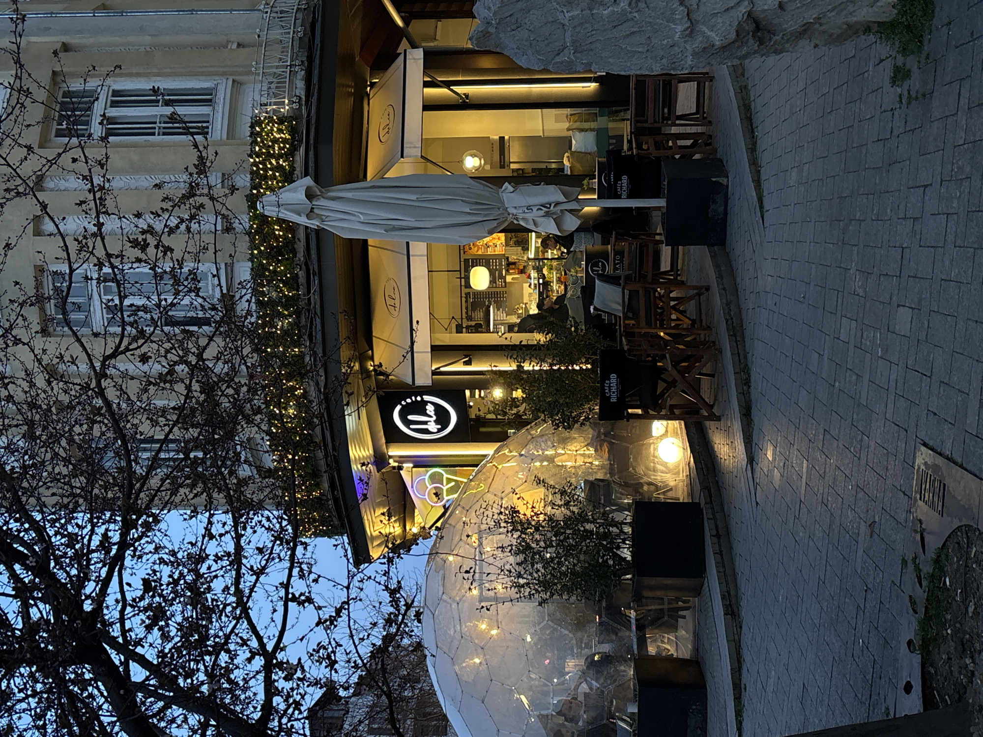 An evening scene depicts a street corner in Varna, Bulgaria, featuring "Dolce cafe." The cafe occupies a ground-floor and corner unit of a multi-story, light-colored building with classical architectural details and windows. The sky is dark blue, indicating dusk or night.
The cafe facade is brightly illuminated with internal lights, a circular white neon sign displaying "Dolce cafe" and an ice cream cone graphic, and external spot lighting. White awnings extend over the outdoor seating area directly in front of the cafe. A large, light-colored patio umbrella is folded and positioned centrally in this area. Wooden tables and chairs are arranged both directly outside the cafe and within a large, transparent, multi-faceted geodesic dome structure positioned to the left of the main cafe entrance. This dome is also internally illuminated with warm lights, and multiple individuals are visible seated inside. Through the cafe's glass windows, additional individuals are discernible inside near a service counter.
Above the cafe, a balcony railing is adorned with green foliage and numerous small, glowing string lights. A large deciduous tree with bare branches partially obscures the upper left portion of the building and the sky. The ground consists of dark grey rectangular pavers. Potted plants are strategically placed around the outdoor seating and within the dome. Additionally, glowing spherical lights are visible on the ground near the dome. A dark sign displaying "CAFES RICHARD" in white text is visible near the outdoor seating. On the far right, a large, irregularly shaped natural rock formation is partially visible. A metal grate and disturbed earth are present in the lower-left foreground.