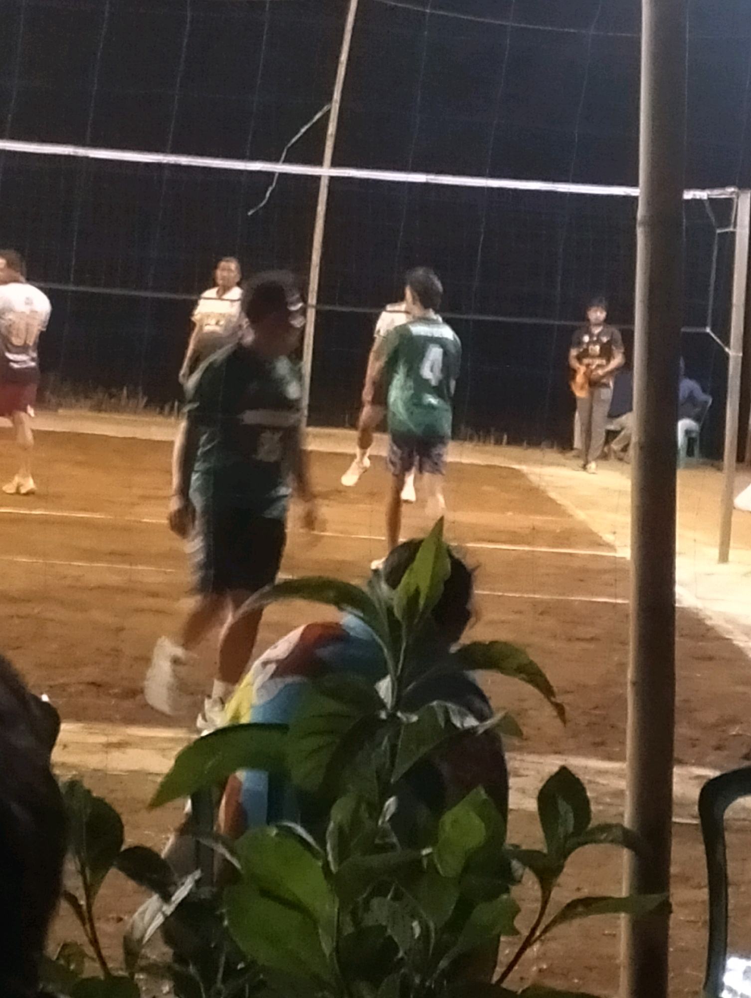 In Krondonan, Indonesia, a volleyball game is underway on a dirt court under artificial lighting. A volleyball net divides the court, with a visible white line marking the boundary. Several individuals are present, appearing to be players and possibly spectators. One player in the foreground wears a dark green jersey with the number "4" on the back. Other individuals are seen in athletic attire, some with their backs to the camera. The environment is dimly lit, suggesting an evening or nighttime setting. The ground is primarily composed of dirt, with some sparse vegetation visible in the foreground. The scene captures a moment during a casual volleyball match.