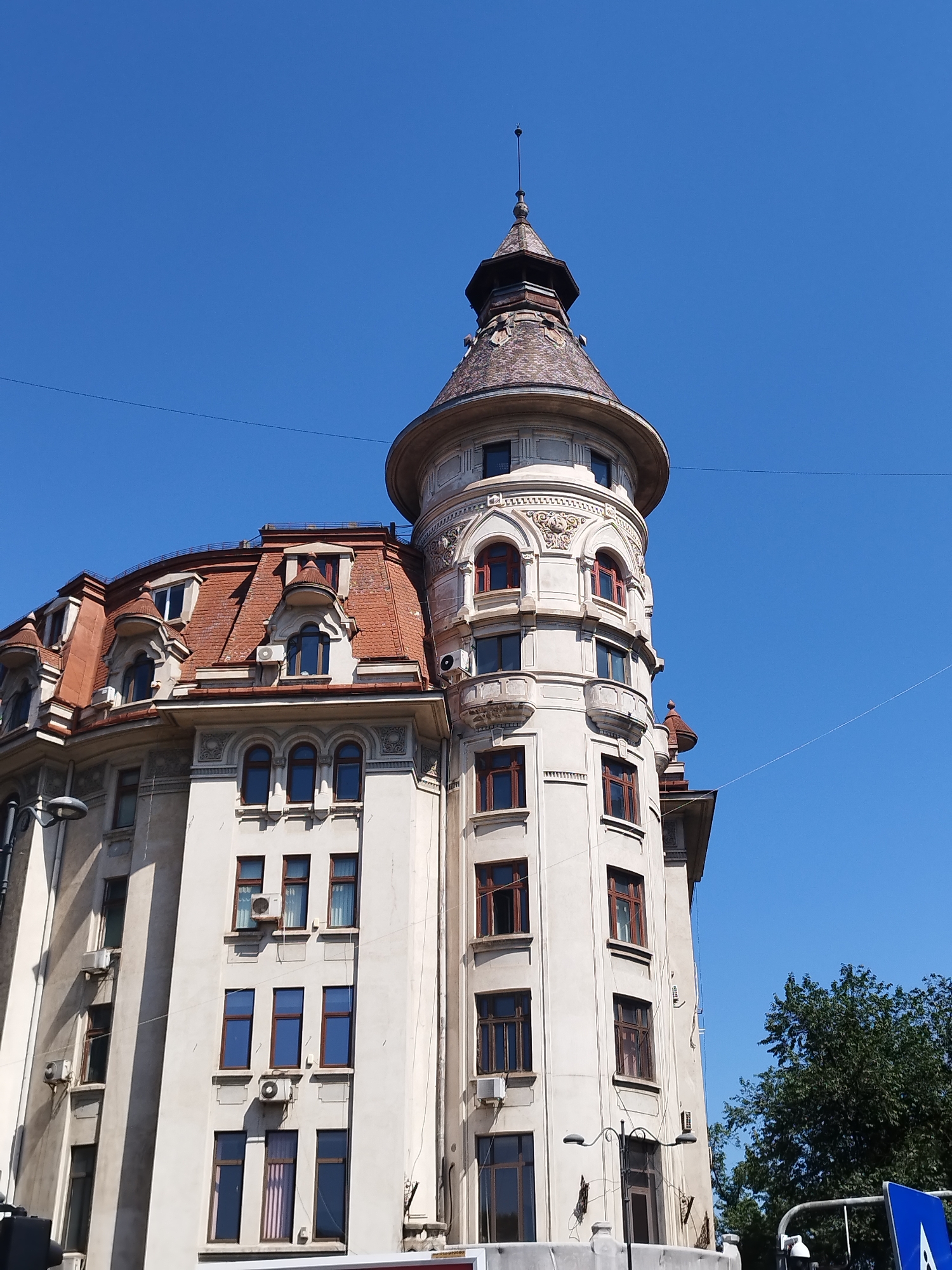 The image captures the exterior of a building with a distinct turret and mansard roof under a clear blue sky in Bucharest, Romania. The building appears to be made of light-colored stucco and features numerous windows with dark frames, some of which are arched. The turret is topped with a conical roof covered in tiles, and a thin spire extends from its peak. The mansard roof also has red tiles and several dormer windows. Green trees are visible on the right side of the image, and overhead wires are present against the sky. The architectural style suggests it is an older building, likely from the late 19th or early 20th century. There are no people visible in the frame.