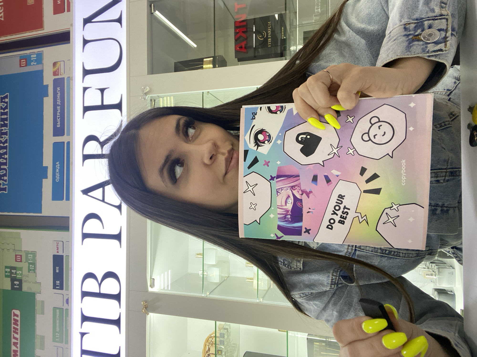 The image shows a young woman holding a notebook with "DO YOUR BEST" printed on it, positioned in front of a store display in a shopping mall. A sign above reads "PARFUM." The woman has long dark hair and is wearing a denim jacket. Her nails are painted bright yellow. Behind her is a store display case with perfume bottles. A directory map to the left indicates sections such as "MAGNET," "ADMINISTRATION," and "FAST MONEY." The directory also features Russian text. Based on the OCR'd text "PARFUM" and the directory including "MAGNET" and other indications, this image appears to be taken in a retail environment. Specifically, based on the text on the directory, it appears that this location is in the city of Chaikovsky, Russia.