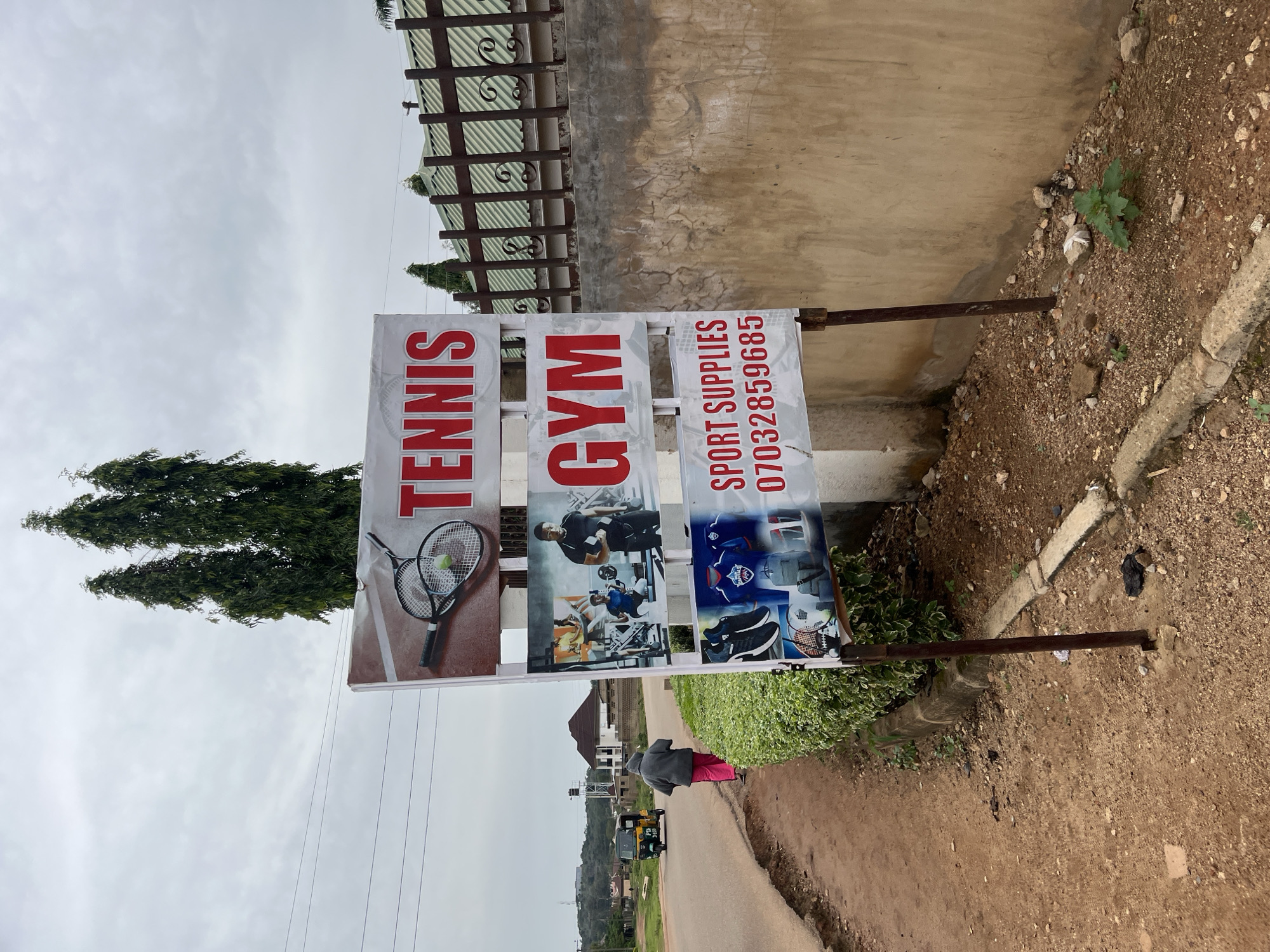 A street-level, outdoor, medium shot captures a sign for "TENNIS GYM SPORT SUPPLIES" in Bukuru, Nigeria. The sign, mounted on a metal pole, is comprised of several panels. The top panel displays a tennis racket and ball. Below this, the words "TENNIS GYM" are prominently displayed in large red letters. The bottom panel reads "SPORT SUPPLIES" and a phone number, "07032859585". Images of athletic wear and equipment are also visible on the lower portion of the sign.
The sign stands beside a dirt and gravel roadside, lined with a low, manicured green hedge. To the left of the sign, a person in a grey hooded jacket and pink trousers walks away from the camera along the paved road. A yellow and green auto-rickshaw is visible further down the road. In the background, several tall, slender trees rise above a brown-tiled roof and a metal fence. The sky is overcast with a few scattered clouds. The scene is illuminated by ambient daylight.