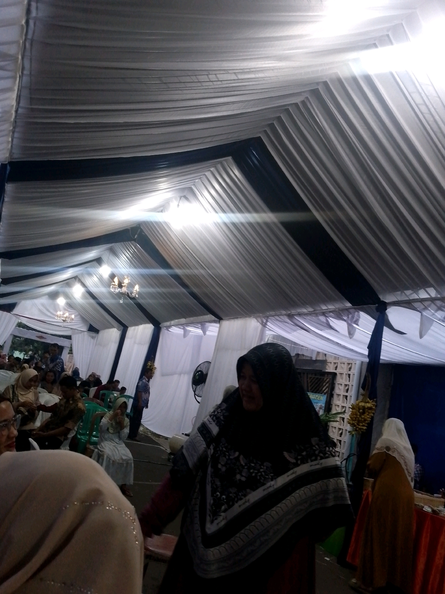 The image depicts an indoor event tent, likely a celebration or gathering, in Tigaraksa, Indonesia. The tent's interior is draped with white and dark blue fabric, forming a ribbed ceiling structure illuminated by chandeliers and spotlights. The scene shows numerous people, many of whom are women wearing hijabs, seated or standing within the tent. Some individuals are engaged in conversation, while others appear to be observing the event. The foreground features a woman in a black hijab and a patterned scarf, partially obscuring her face. To the right, another woman in a white hijab and a yellow dress stands near a table covered with an orange tablecloth. In the background, there are more attendees, chairs, and a fan. The overall atmosphere suggests a communal gathering or ceremony.