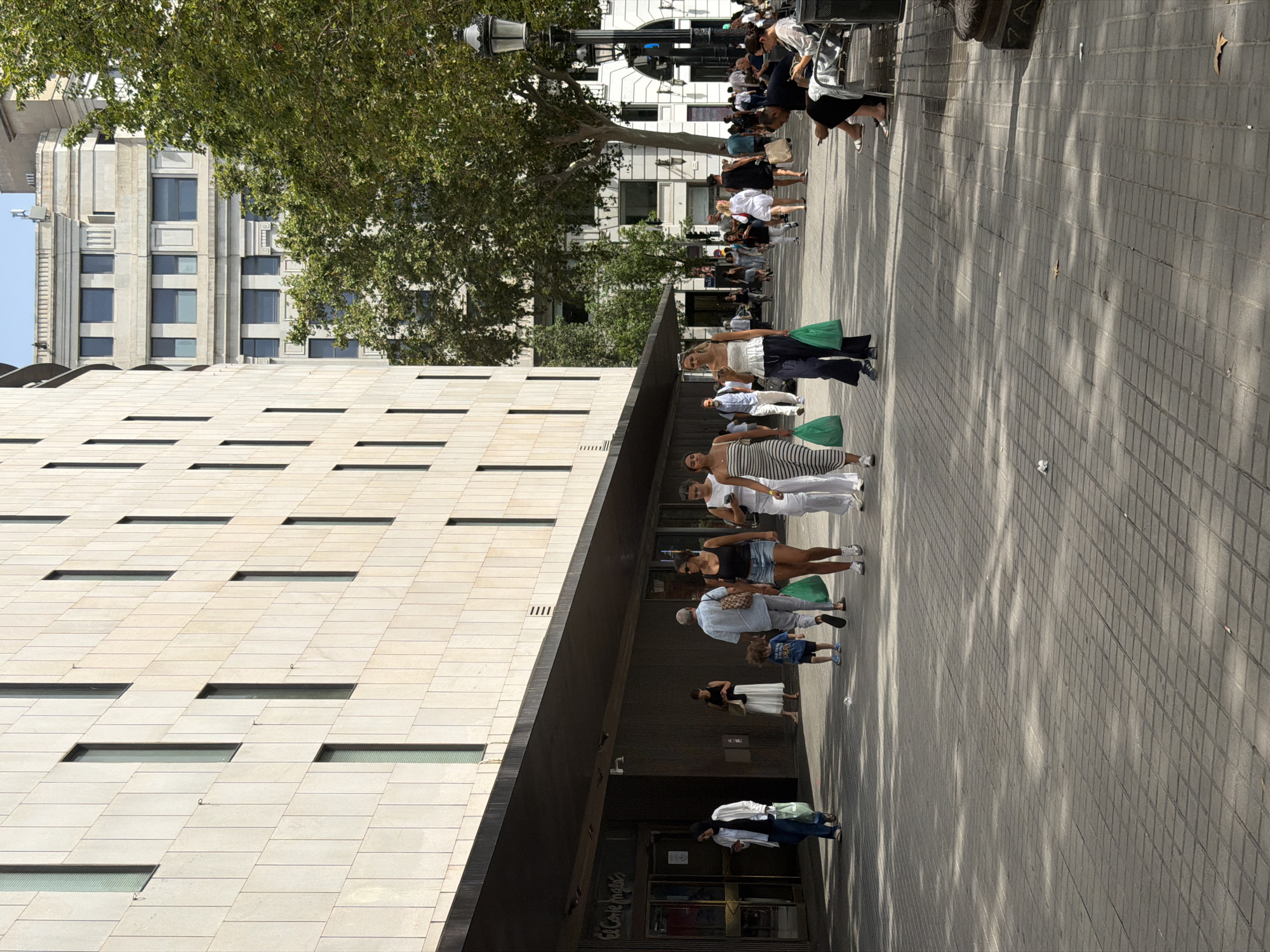 The image shows a street scene in Barcelona, Spain, in front of El Corte Inglés department store. A group of people are walking on the tiled sidewalk. On the left, a woman wearing a hijab and white jacket walks with a shopping bag. To her right, a man and woman with a young child walk past the entrance. Further down the street, several people are seen walking and sitting on benches. A tall building with many windows stands behind the people, and trees line the sidewalk. The sun is shining, casting shadows on the ground.