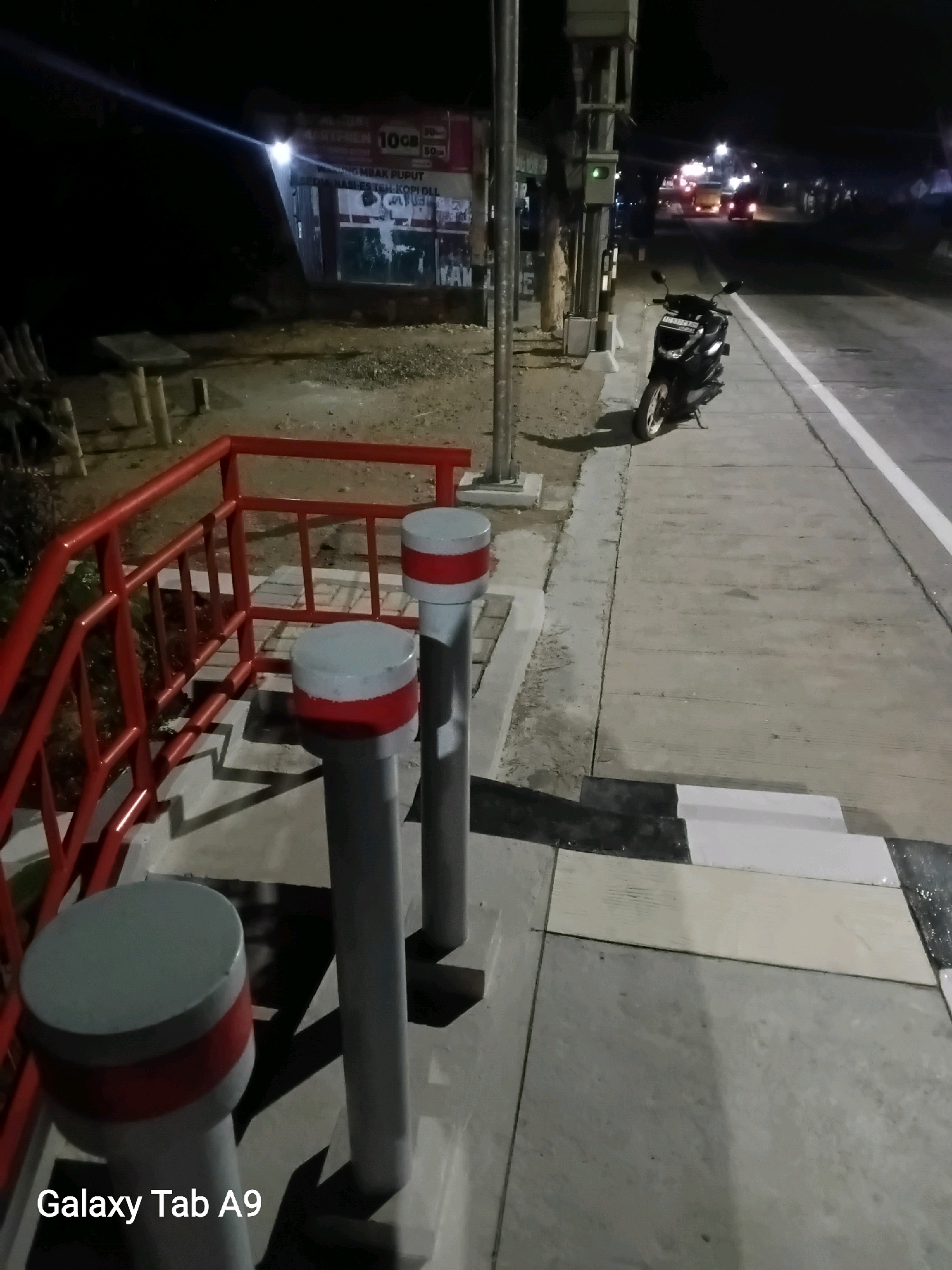 The image shows a street scene at night in Bendo, Indonesia. A black scooter is parked on the sidewalk next to a road. A red railing and several bollards are visible in the foreground. In the background, there are lights from buildings and vehicles on the road, suggesting a commercial or residential area. The overall atmosphere is dark and quiet.