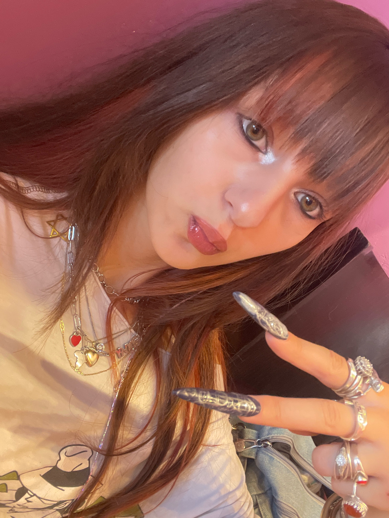 The image is a close-up, selfie-style portrait of a young woman with long, brown hair with reddish highlights and bangs. She is wearing multiple silver necklaces, one with a Star of David pendant, a red heart charm, and a gold heart charm. She has on several silver rings, including a large one on her right index finger, and her fingernails are long and appear to be painted with a silver or dark metallic color. The woman is wearing makeup, including dark eyeliner and lip gloss, and her eyes are looking towards the camera. She is wearing a light-colored t-shirt with a graphic print. The background is a pink wall and a dark surface that may be furniture or an object. The image does not contain any information to indicate a location in Raanana, Israel.