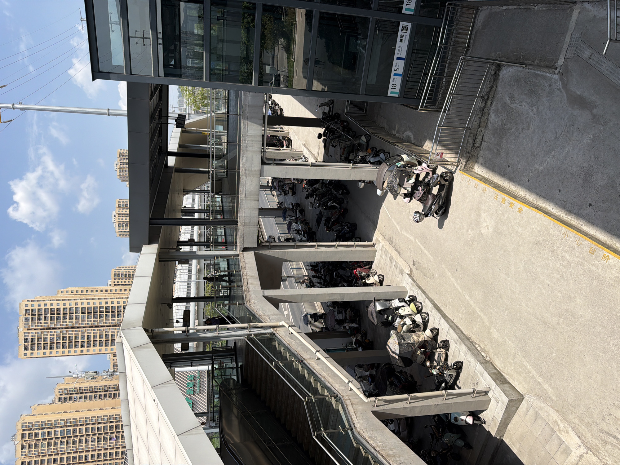 The image depicts an outdoor view of a pedestrian overpass and a surrounding area with parked electric scooters. The overpass, made of concrete and metal, has a sloped ramp and stairs leading up to an elevated walkway with glass railings. Above the walkway, a dark grey canopy extends across. In the background, tall, beige apartment buildings with many windows are visible under a bright blue sky with scattered white clouds. A tall electrical tower with wires stretches upwards.
The ground level below the overpass is a paved area filled with numerous parked electric scooters, some with protective covers. Several people are present: one individual walks on the elevated walkway, another is near the stairs, and a third person is seen on the ground level walking among the scooters. Signs with Chinese characters and numbers like "18号线" (Line 18) and "5号线" (Line 5) are affixed to a glass-enclosed structure on the right side of the image, indicating a public transportation hub, likely a metro station. The area appears to be a section of the city of Tiaodeng, China, given the station names. The overall scene is one of urban infrastructure and daily life, with a focus on transit and personal mobility.