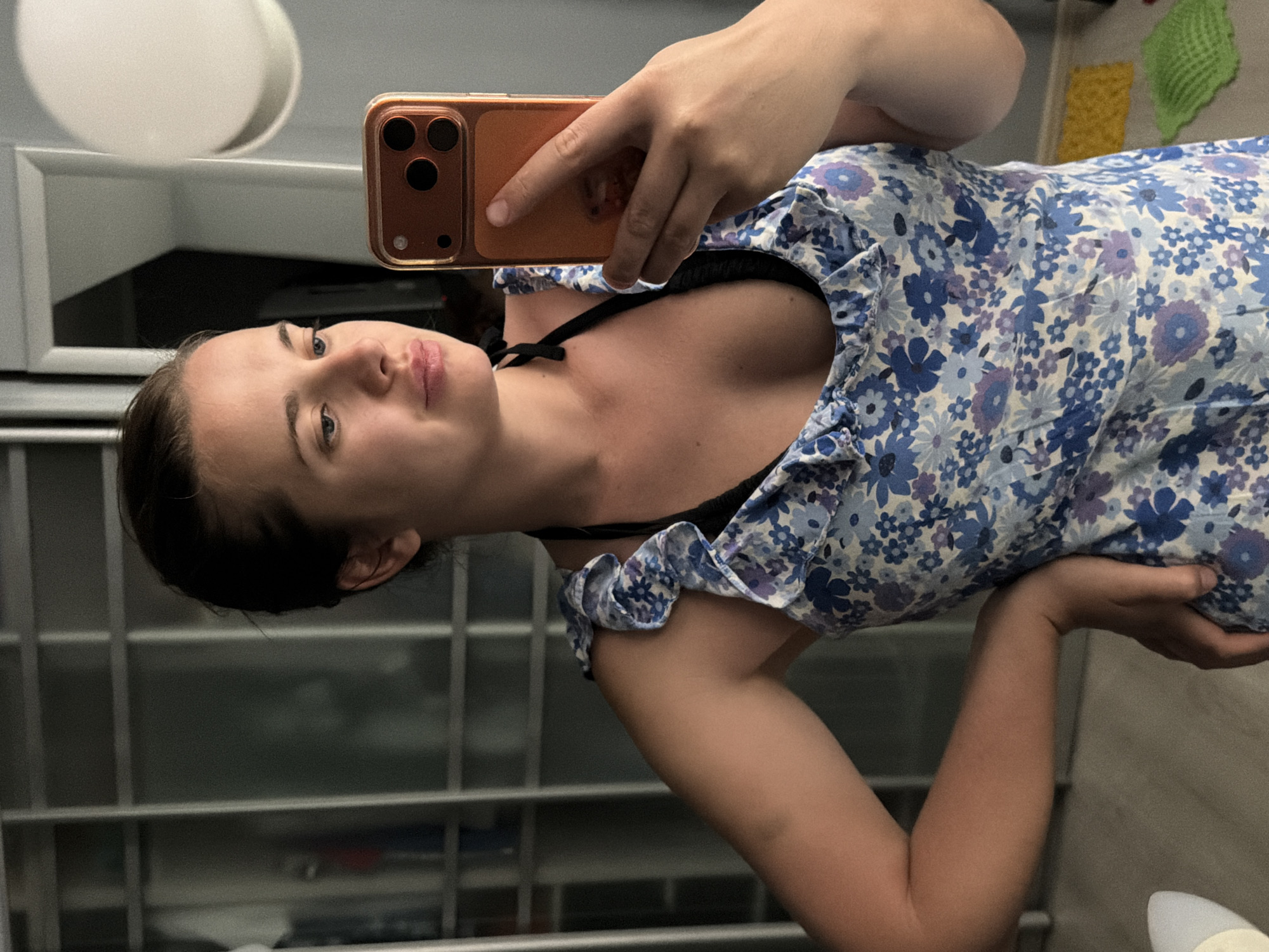 An adult female, estimated 20-30 years of age, with dark hair pulled back, is depicted taking a self-portrait using a mobile phone held in her right hand. She is gazing slightly downwards and to the right, with a neutral expression. Her left arm is bent, with her hand resting on her hip. She wears a blue and white floral patterned dress with ruffled shoulder straps, layered over a visible black bra or swimsuit top.
The smartphone appears to be an iPhone model, encased in a translucent orange/amber cover, clearly showing the triple-lens camera array on the rear. The setting is an indoor domestic environment. The background on the left consists of frosted glass or mirrored sliding wardrobe doors with a white grid frame. To the upper right, a circular white wall-mounted light fixture is prominently visible. The floor is light-colored, and in the lower right corner, two small, textured mats or cloths, one yellow and one green, are present. The scene is located in Sochi, Russia.