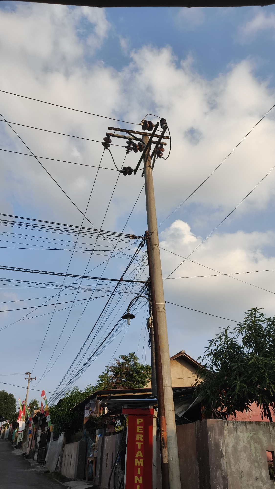 The image depicts a residential street in Bandar Lampung, Indonesia, under a partly cloudy sky. A tall concrete utility pole, laden with numerous electrical wires and insulators, dominates the center of the frame. The pole shows signs of age and weathering. To the right of the pole, a pink-colored wall encloses a property with a large, leafy green tree. Behind the wall, the roofline of a house is visible. In the foreground on the right, a vertical red sign with yellow lettering that reads "PERTAMINI" is partially visible, indicating a small, roadside gas station or convenience store.
To the left of the utility pole, a narrow paved street recedes into the distance. Along the left side of the street, a row of houses and structures are visible, many adorned with flags. The buildings appear to be of various sizes and construction, with awnings and covered areas. Dense foliage is present behind these structures. The overall impression is that of a typical urban street scene in Southeast Asia, characterized by overhead utility lines and a blend of residential and commercial elements. No people are visible in the image.
